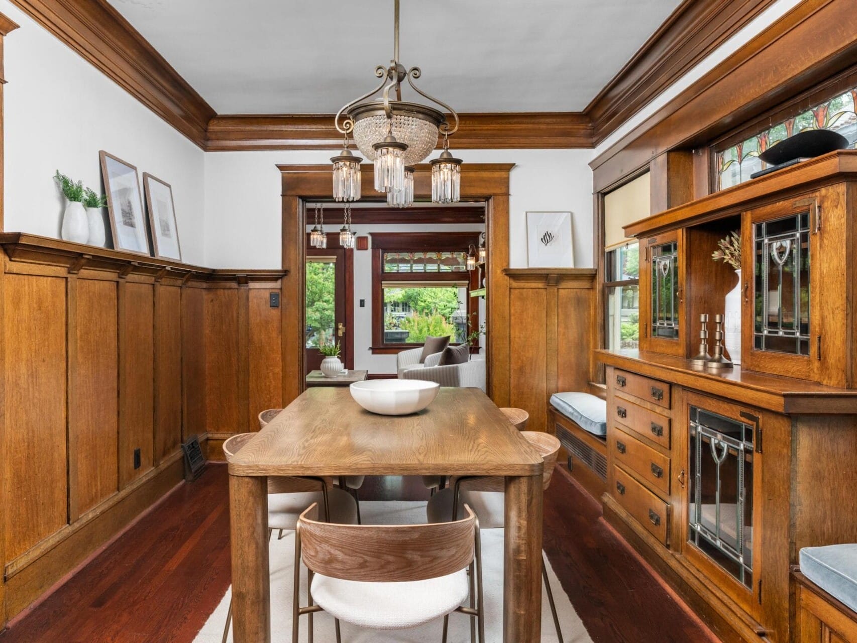 A dining room with wooden paneling and built-in cabinets, featuring a wooden table with four chairs, a modern chandelier, large windows, and decorative items on shelves. Natural light fills the room.