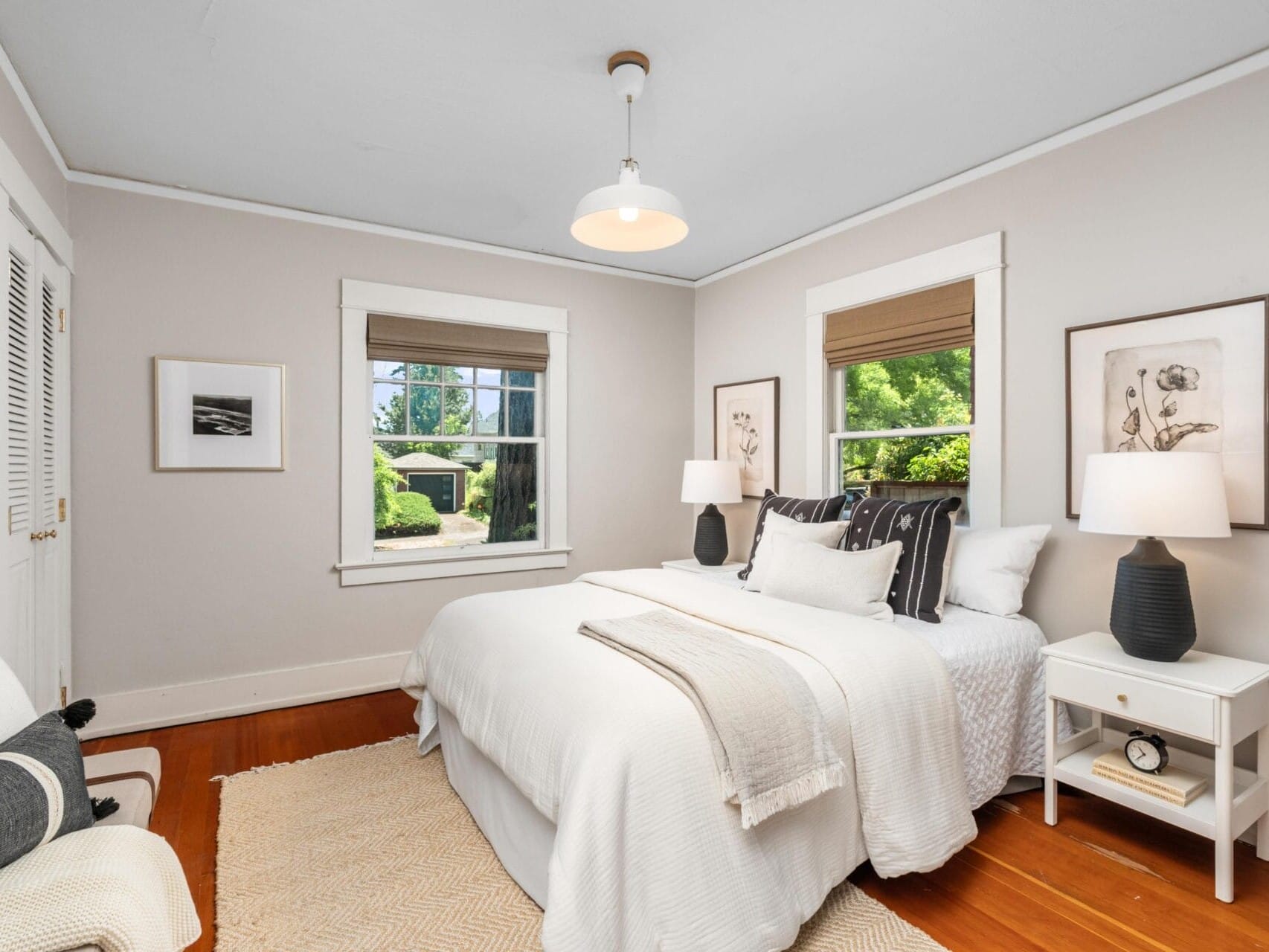 Bright bedroom with a large bed dressed in white bedding, two bedside tables with lamps, framed art on the walls, a window with a garden view, a cushioned chair, and a light fixture hanging from the ceiling.