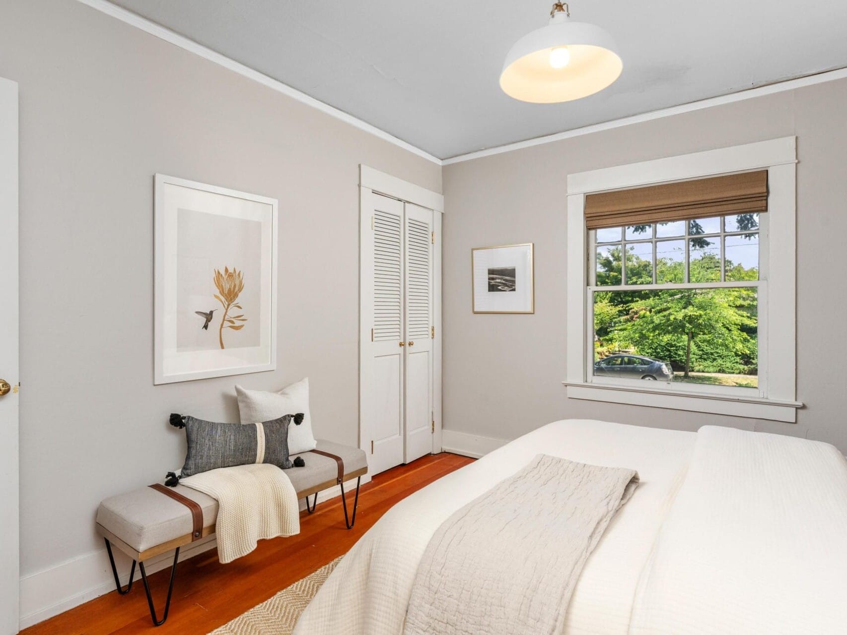A bright, minimalist bedroom with a bed, bench, framed art, and a window showing green trees outside. The room has light walls, wood floors, and a ceiling light fixture.