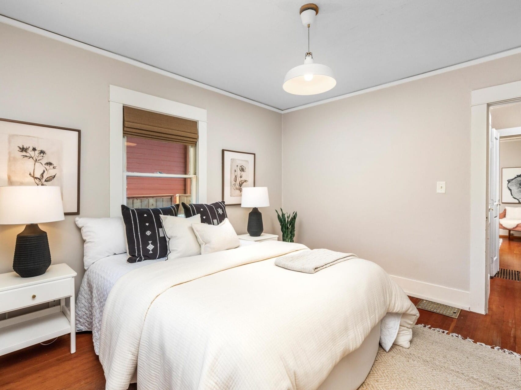 A cozy bedroom with a neatly made bed, white bedding, two bedside tables with lamps, botanical wall art, a window with blinds, a plant in the corner, and warm wooden floors.