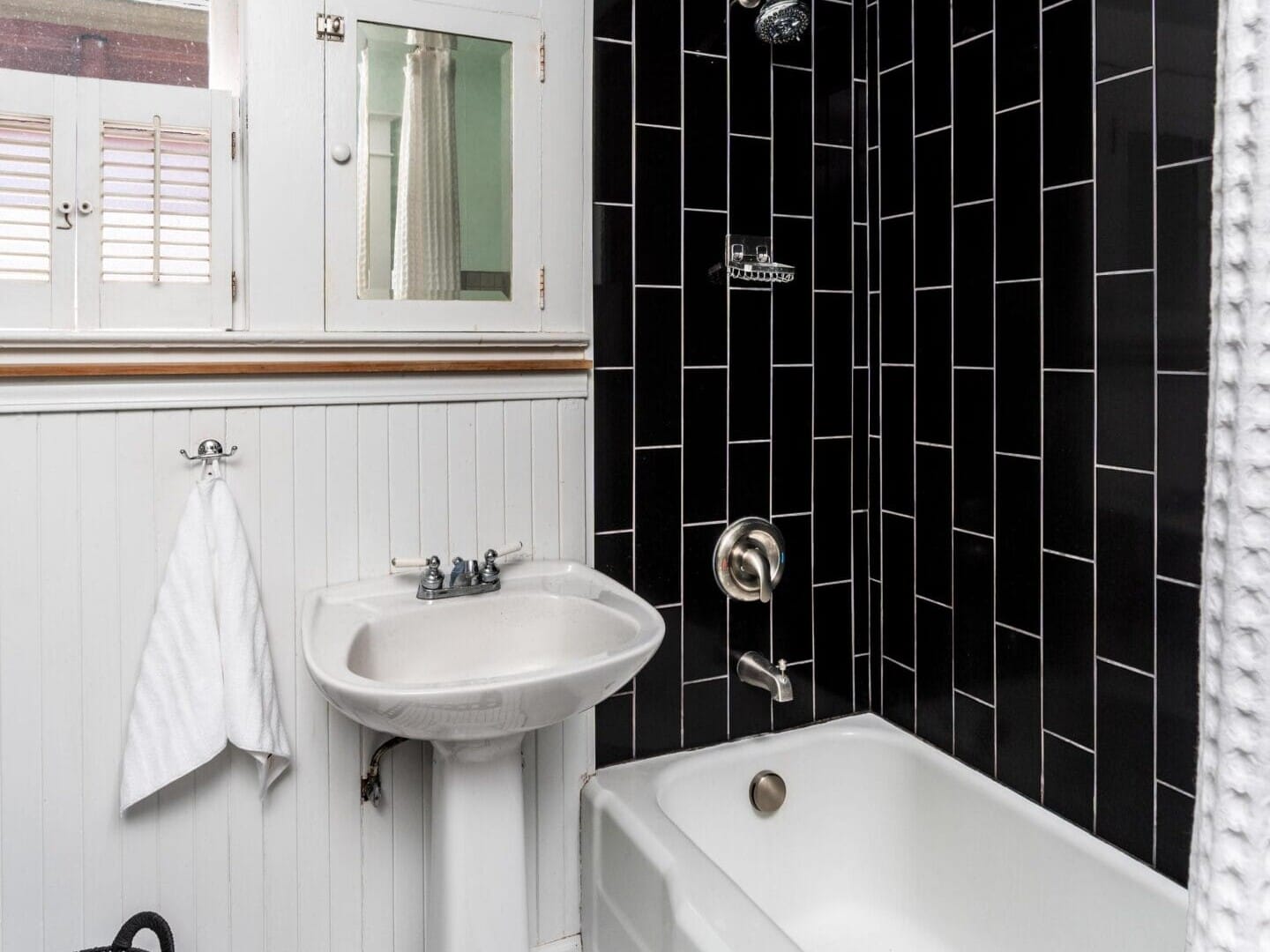 A small bathroom with a white pedestal sink, a bathtub, black vertical tile shower wall, white beadboard wainscoting, mosaic tile floor, a towel on a hook, and a black laundry basket.