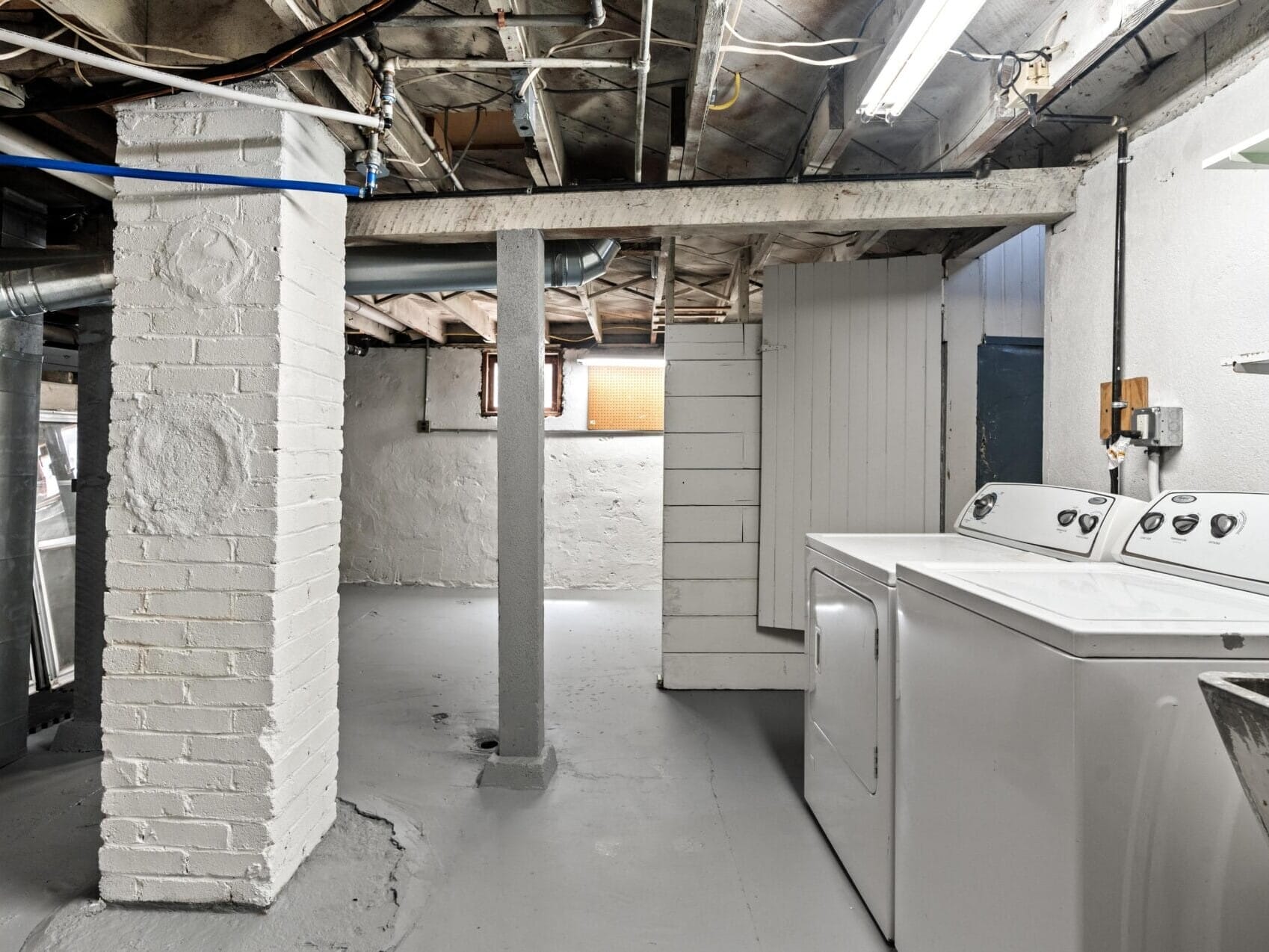 A basement laundry room with exposed ceiling beams, painted white walls and floors, a washer and dryer, shelves with detergent, and a small window letting in light.