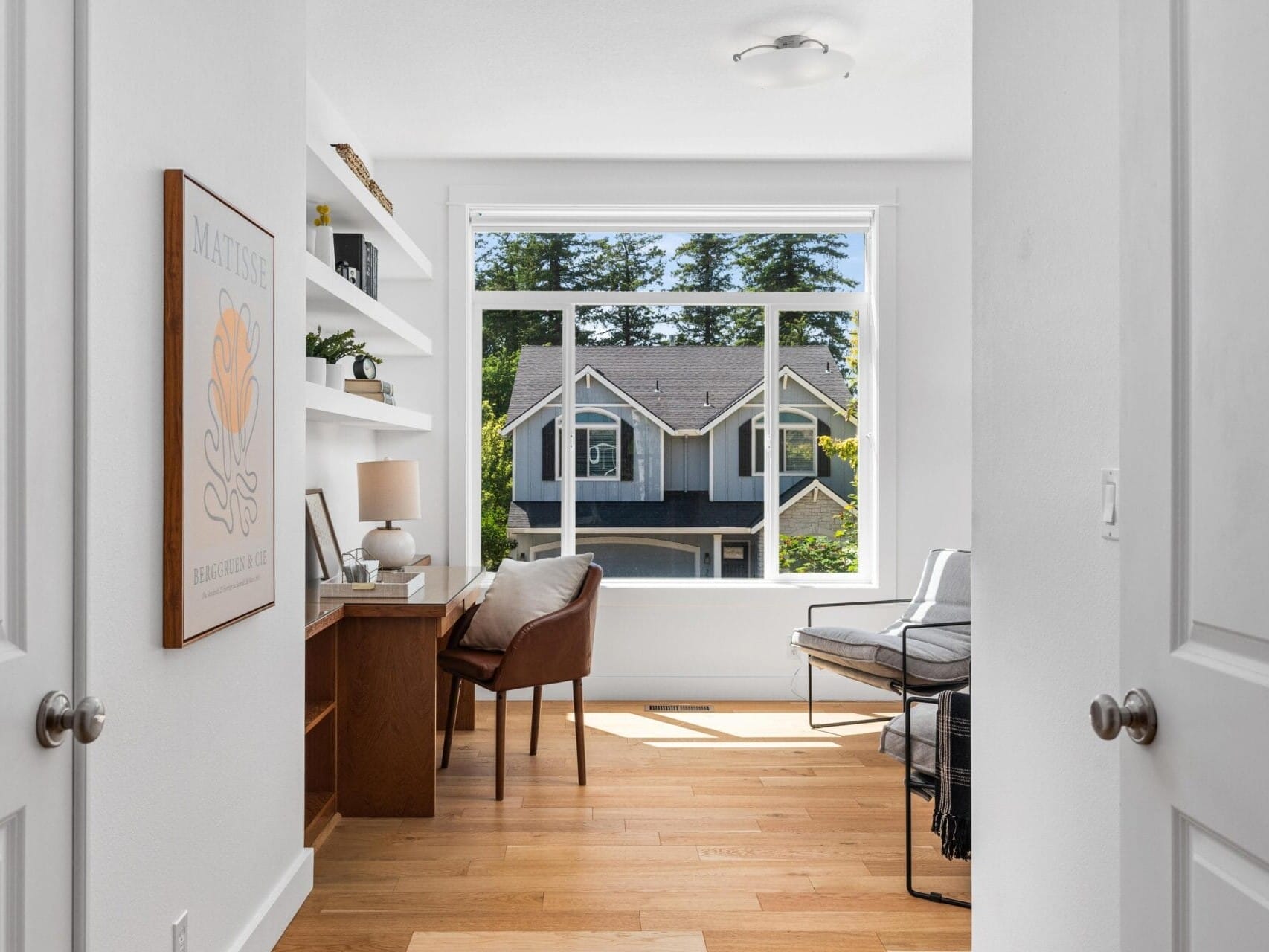 A bright home office with wooden floors, a desk and chair by built-in shelves, a modern lounge chair, and a large window overlooking trees and neighboring houses.