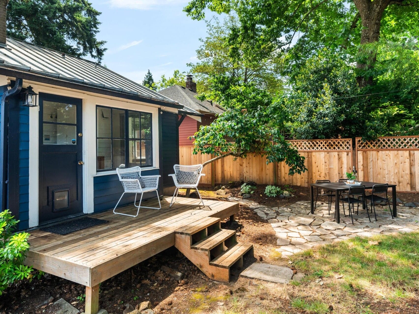 A small blue house with a wooden porch, two white chairs, and a fenced backyard featuring a stone patio with a dining table and four chairs, surrounded by trees and greenery.