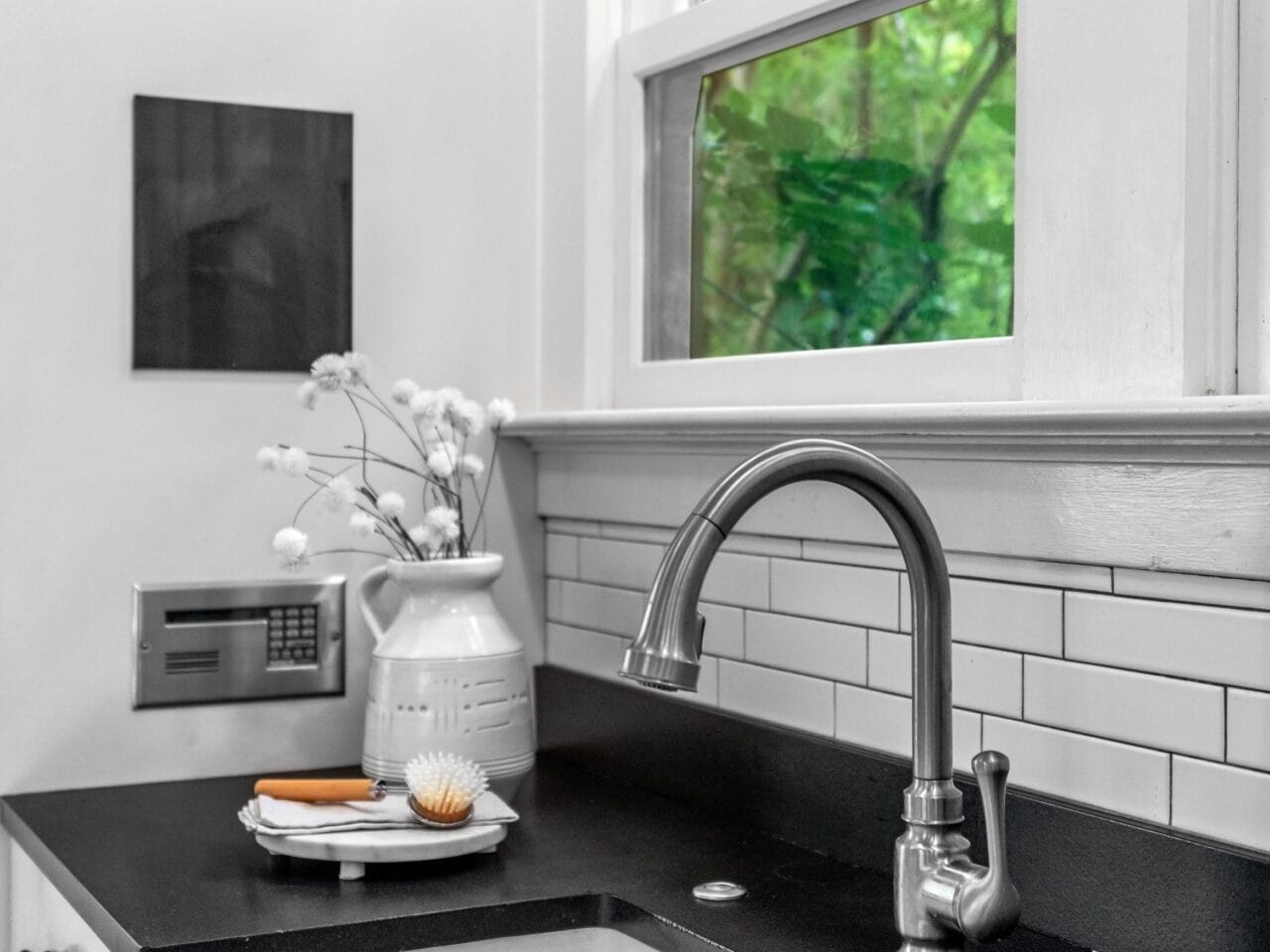 A modern kitchen sink with a stainless steel faucet, black countertop, white farmhouse sink, white subway tile backsplash, and a window showing green trees outside. A vase and dish brush are on the counter.
