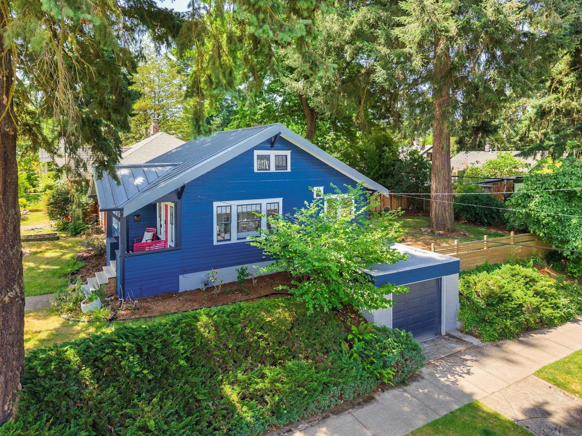 A blue craftsman-style house with a metal roof and a detached garage sits on a tree-lined street, surrounded by greenery and large pine trees. A small porch with red chairs is visible to the left.
