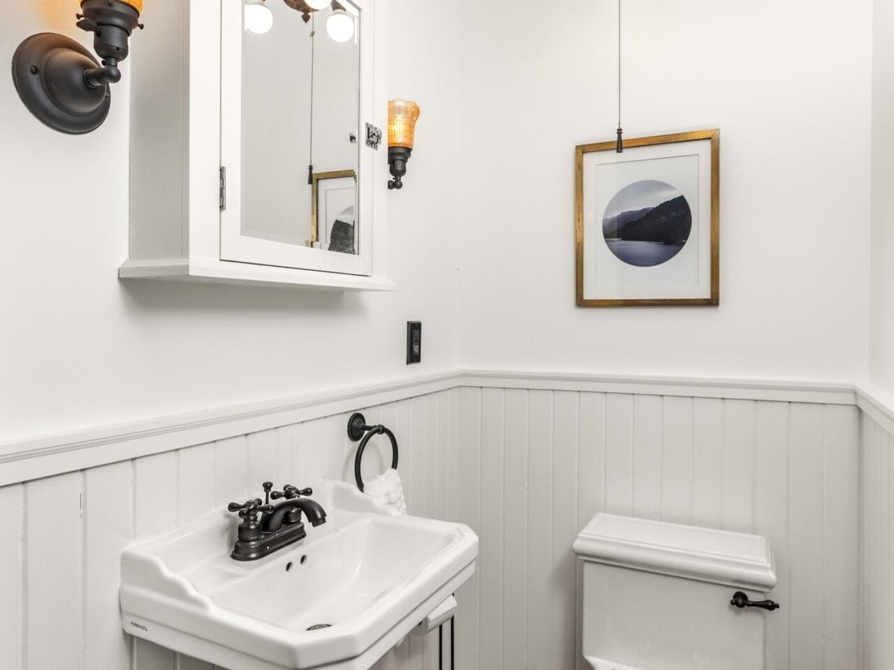 Small bathroom with white wainscoting, a pedestal sink, a toilet with a wooden seat, a wall-mounted mirror cabinet, two vintage-style wall lights, a pendant light, and abstract art on the wall.