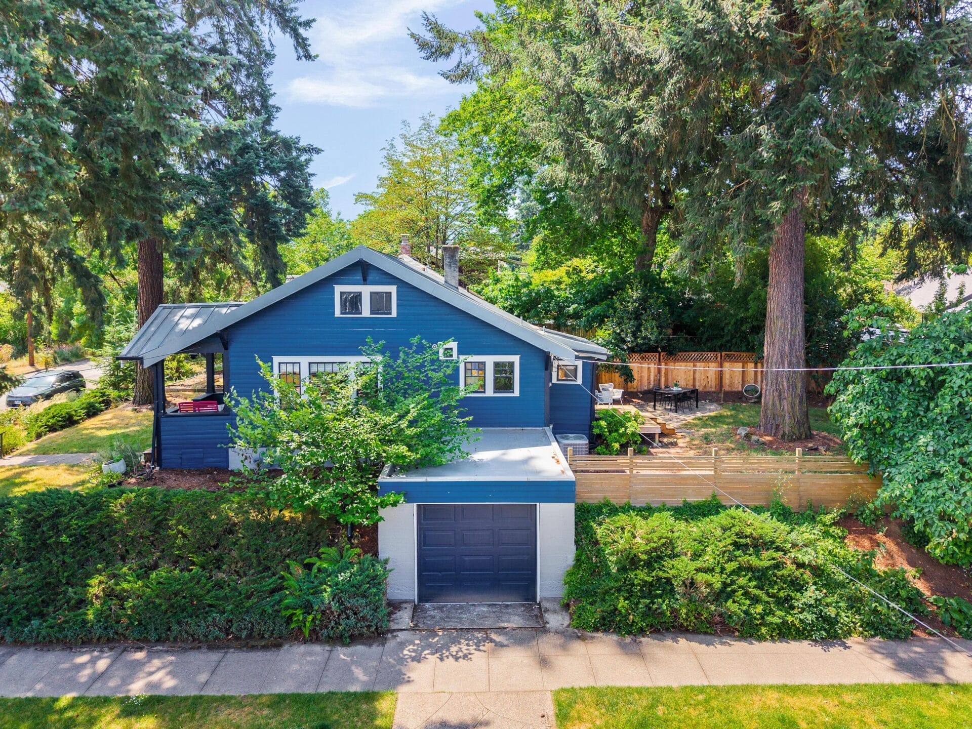 A blue house with a covered porch sits among tall trees and greenery. A garage with a dark blue door is attached, and a fenced backyard is visible on the right. The home is surrounded by lush landscaping and a sidewalk.
