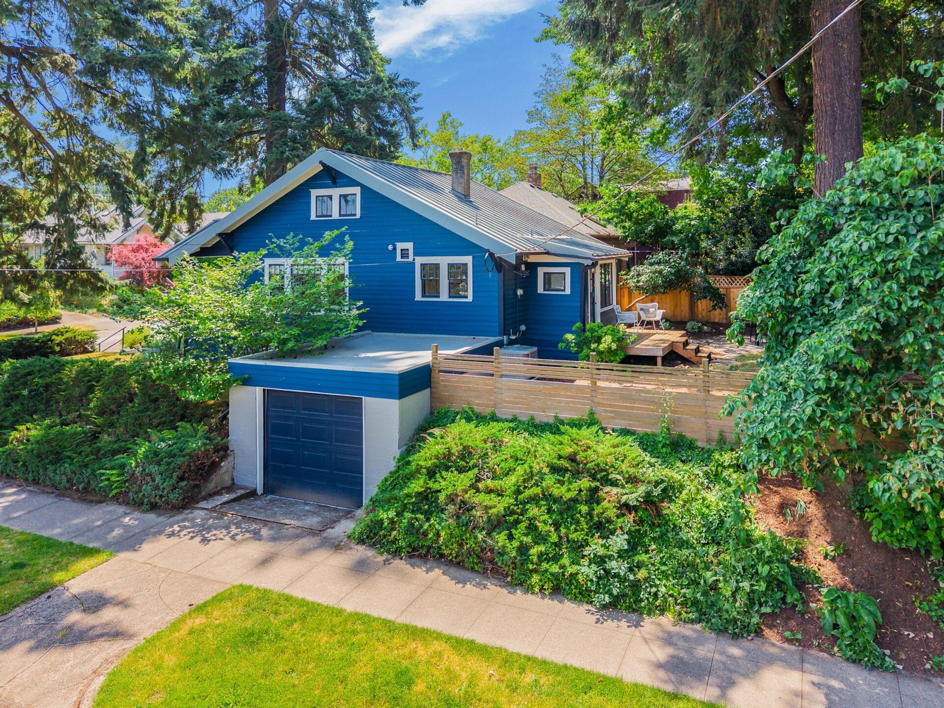 A blue house with white trim sits behind lush green shrubs and trees, featuring a detached blue garage and a wooden fenced patio area, all under a sunny sky.
