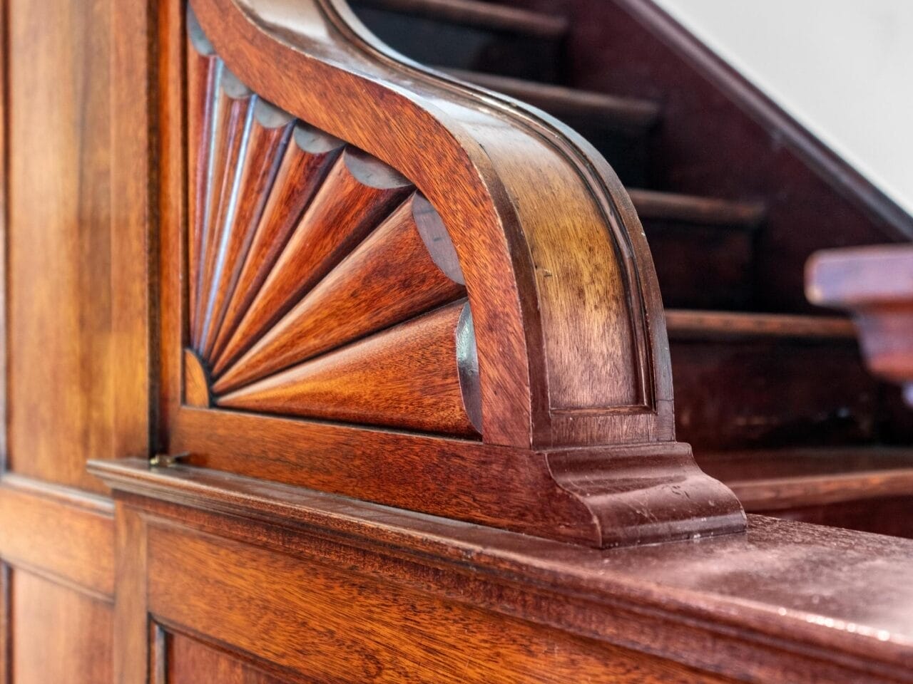 Close-up of a polished wooden staircase banister with an ornate carved detail and glossy finish, showing part of the stairs and a black light switch on the paneled wall.