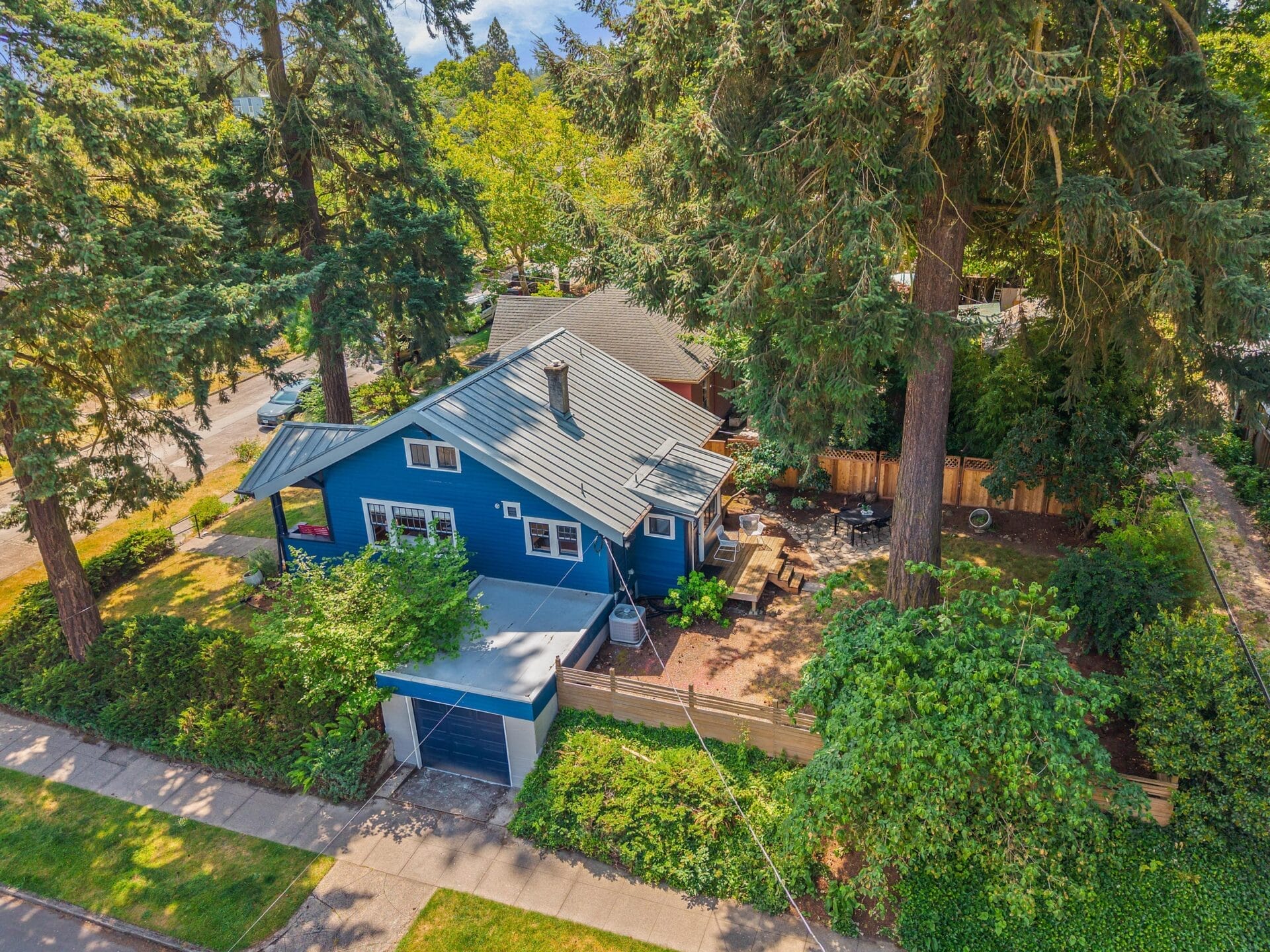 Aerial view of a blue house with a metal roof, surrounded by tall trees and greenery, featuring a front porch, small driveway, and fenced backyard on a residential street.
