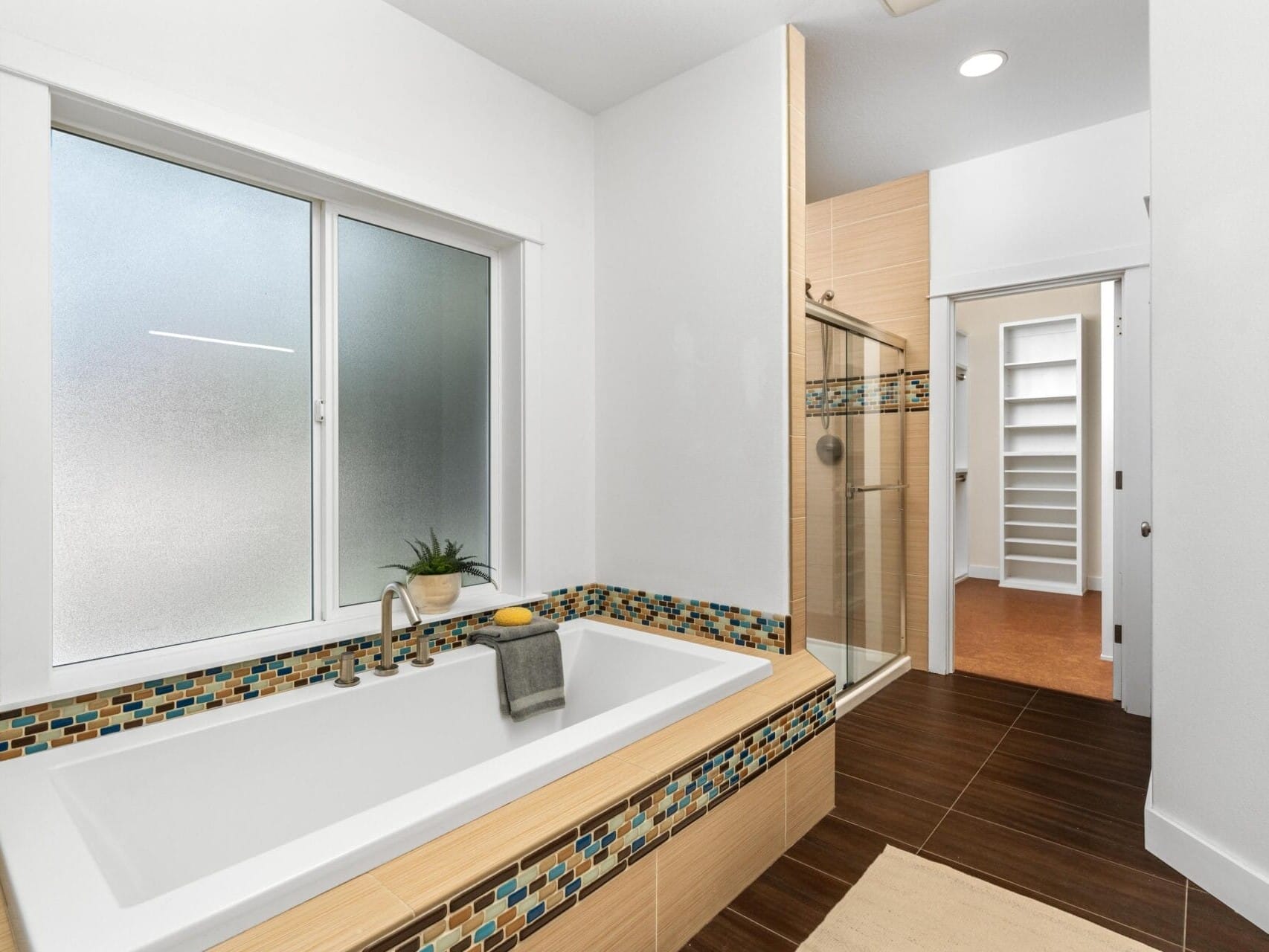Modern bathroom with a white bathtub featuring mosaic tile accents, a glass-enclosed shower, frosted window, towel racks, and a view into a walk-in closet with built-in shelving.