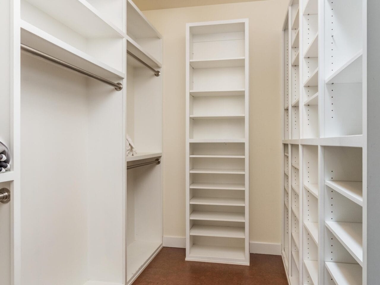 A spacious walk-in closet with white built-in shelves, open hanging rods, and empty storage compartments, featuring a brown floor and a modern ceiling light.