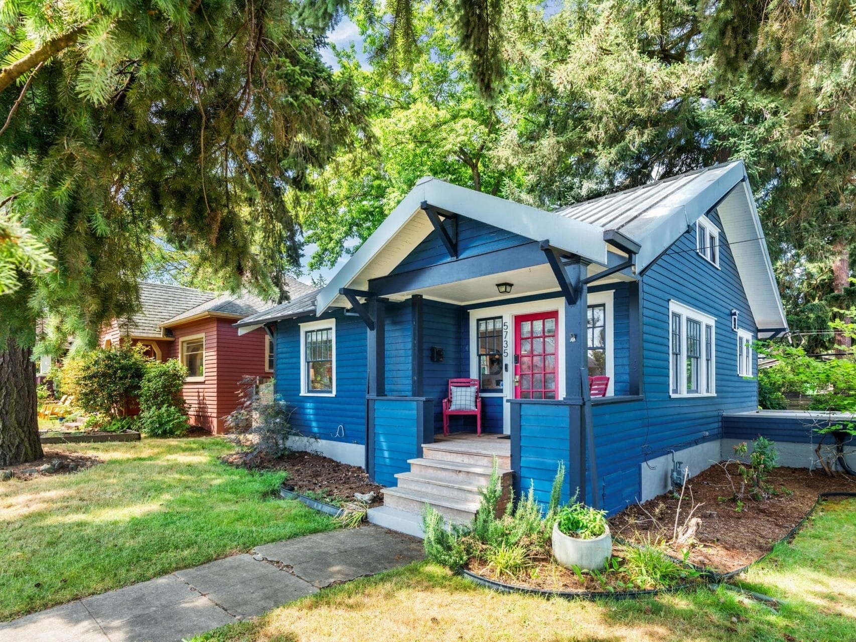 Blue cottage-style house with a covered front porch, white trim, two red chairs, and surrounded by green grass, trees, and shrubs on a sunny day. A neighboring house is partially visible to the left.