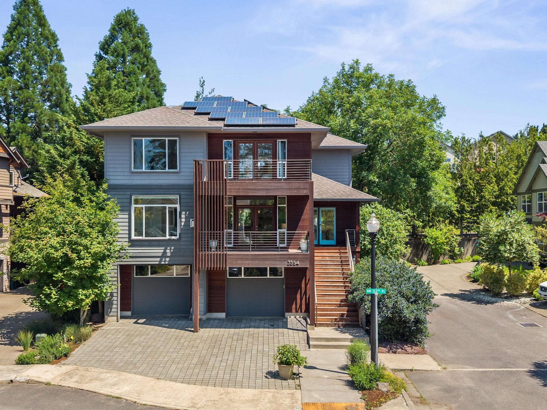 Modern three-story house with solar panels on the roof, large windows, balconies with metal railings, two garage doors, and surrounded by trees on a sunny day at a street corner.
