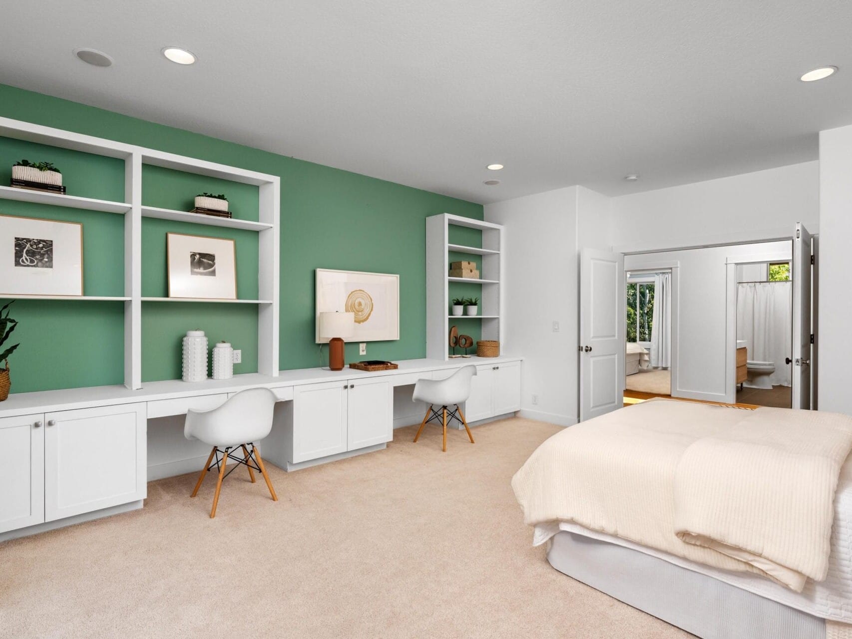 Modern bedroom with a neatly made bed, a green accent wall, built-in white shelves and cabinets, a long desk with two chairs, and decor including plants and framed art. A bathroom is visible through open doors.