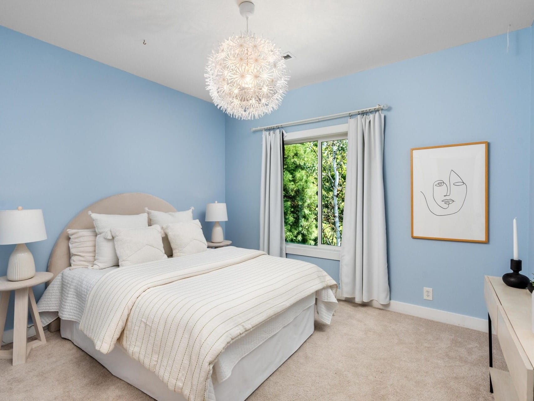 A cozy bedroom with light blue walls, a bed with neutral bedding, two bedside tables with lamps, a window with white curtains, a modern art piece, and a decorative chandelier overhead.