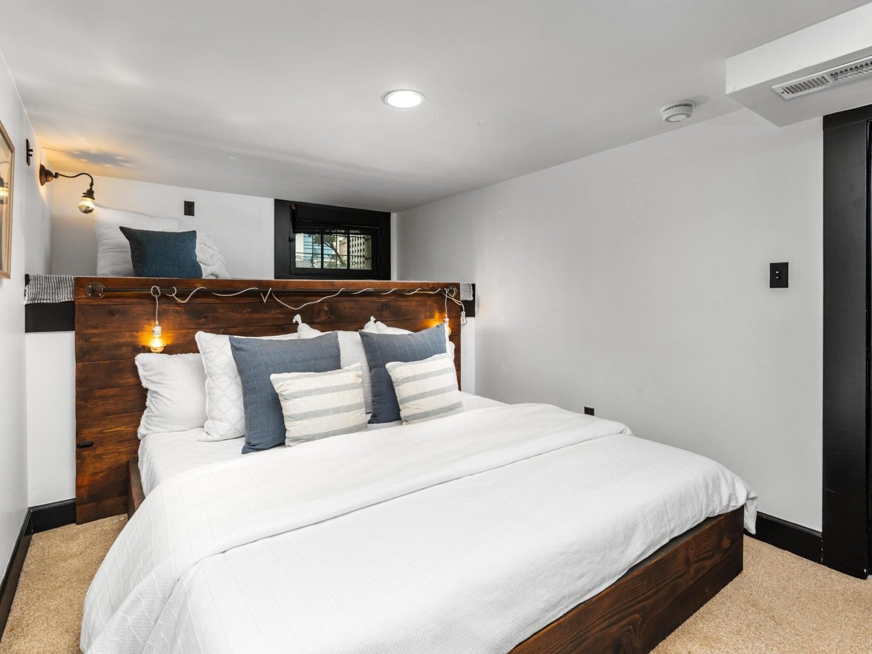 A modern bedroom with a wooden bed frame, white bedding, and multiple white and blue pillows. There are wall-mounted lights above the bed, white walls, beige carpet, and a small window near the ceiling.