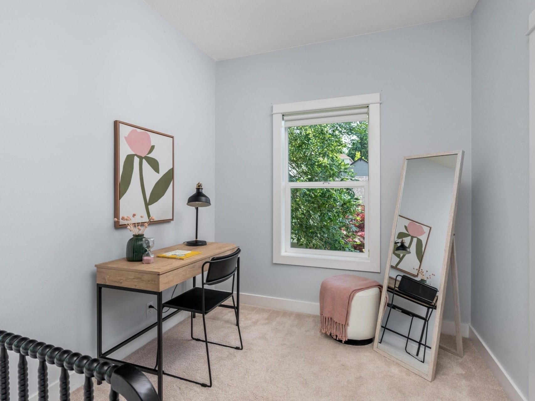 A small home office with a wooden desk, black chair, table lamp, framed flower art, window with leafy tree view, floor mirror, and a white ottoman draped with a pink throw. The walls are light blue and carpet is beige.