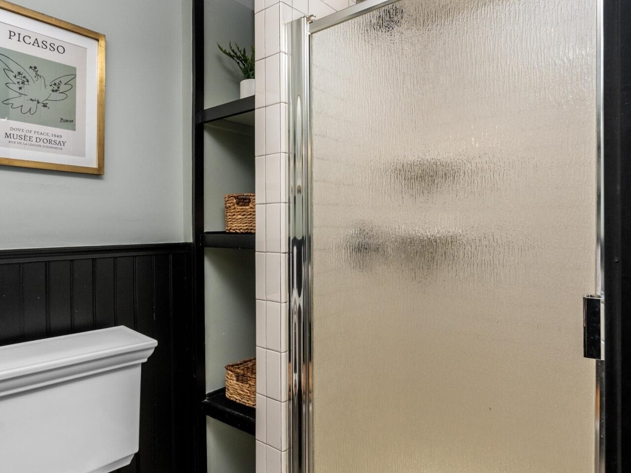 A bathroom with a white toilet featuring a wooden seat, a glass shower with a textured door, black shelving with baskets and a plant, and a framed Picasso poster on a light green wall.