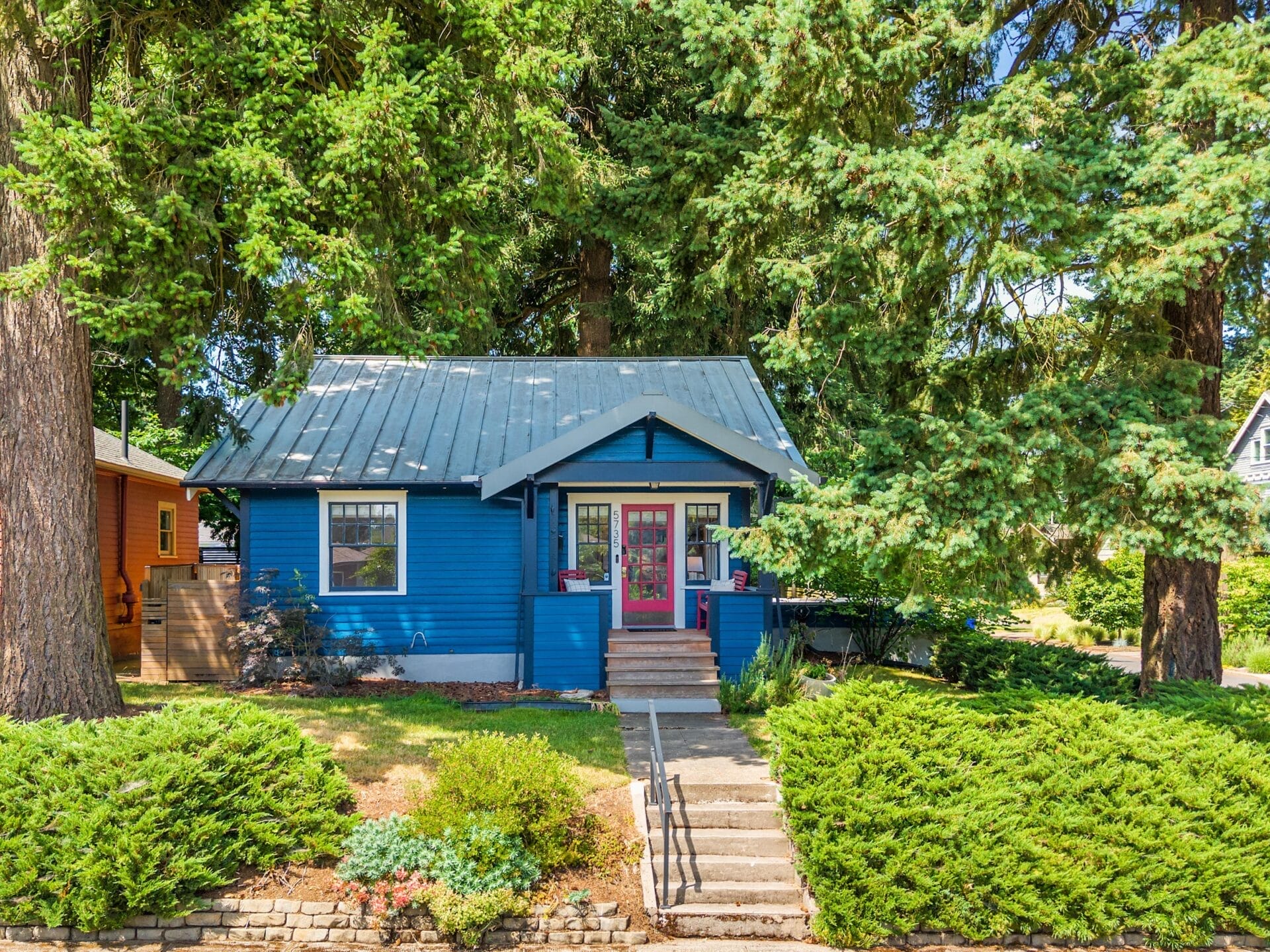 A small blue house with a gray metal roof, red front door, and a front porch, surrounded by large evergreen trees and green shrubs, seen on a sunny day. Steps and a handrail lead up to the entrance.
