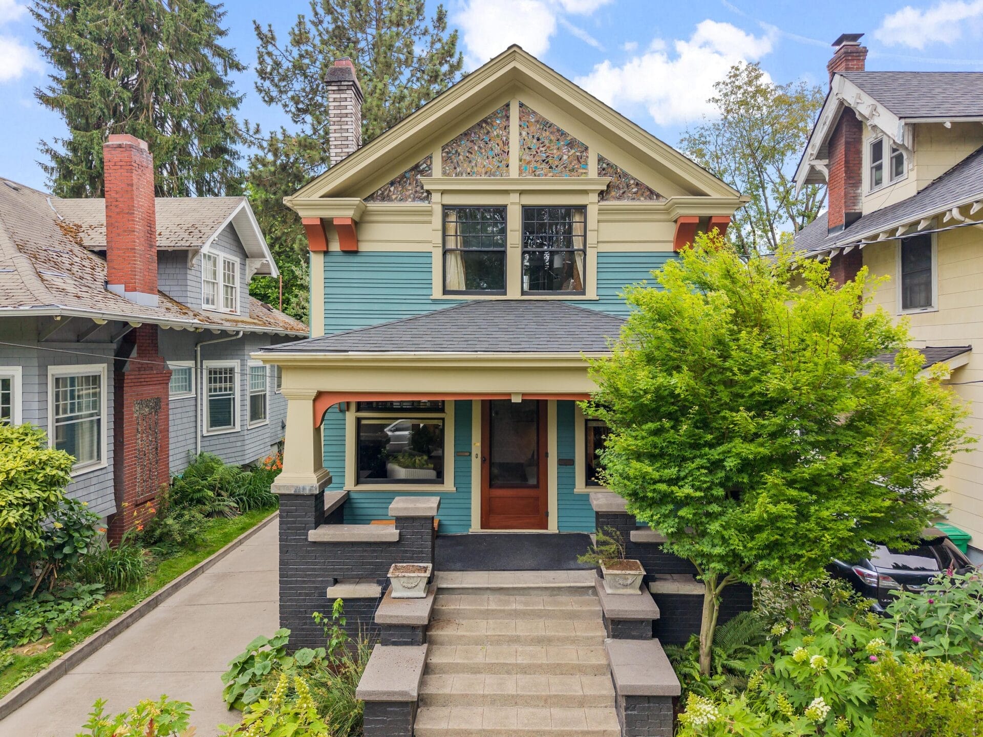 A two-story Craftsman-style house with blue siding, black brick accents, stained glass windows, and a front porch, surrounded by greenery and neighboring houses under a blue sky with clouds.