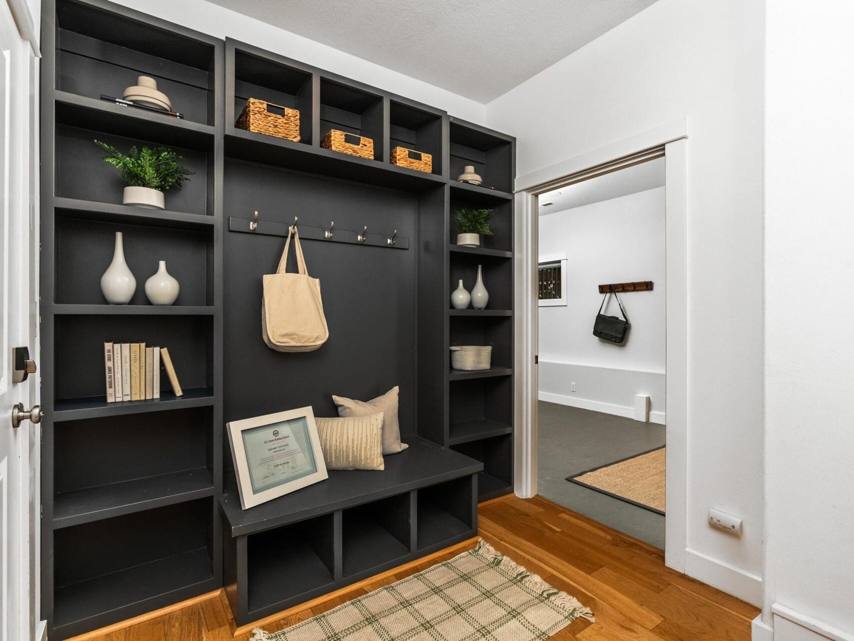 A modern mudroom with black built-in shelves, decorative items, hooks with a hanging tote, a small rug, two pillows, and visible adjacent rooms with white walls and hardwood floors.