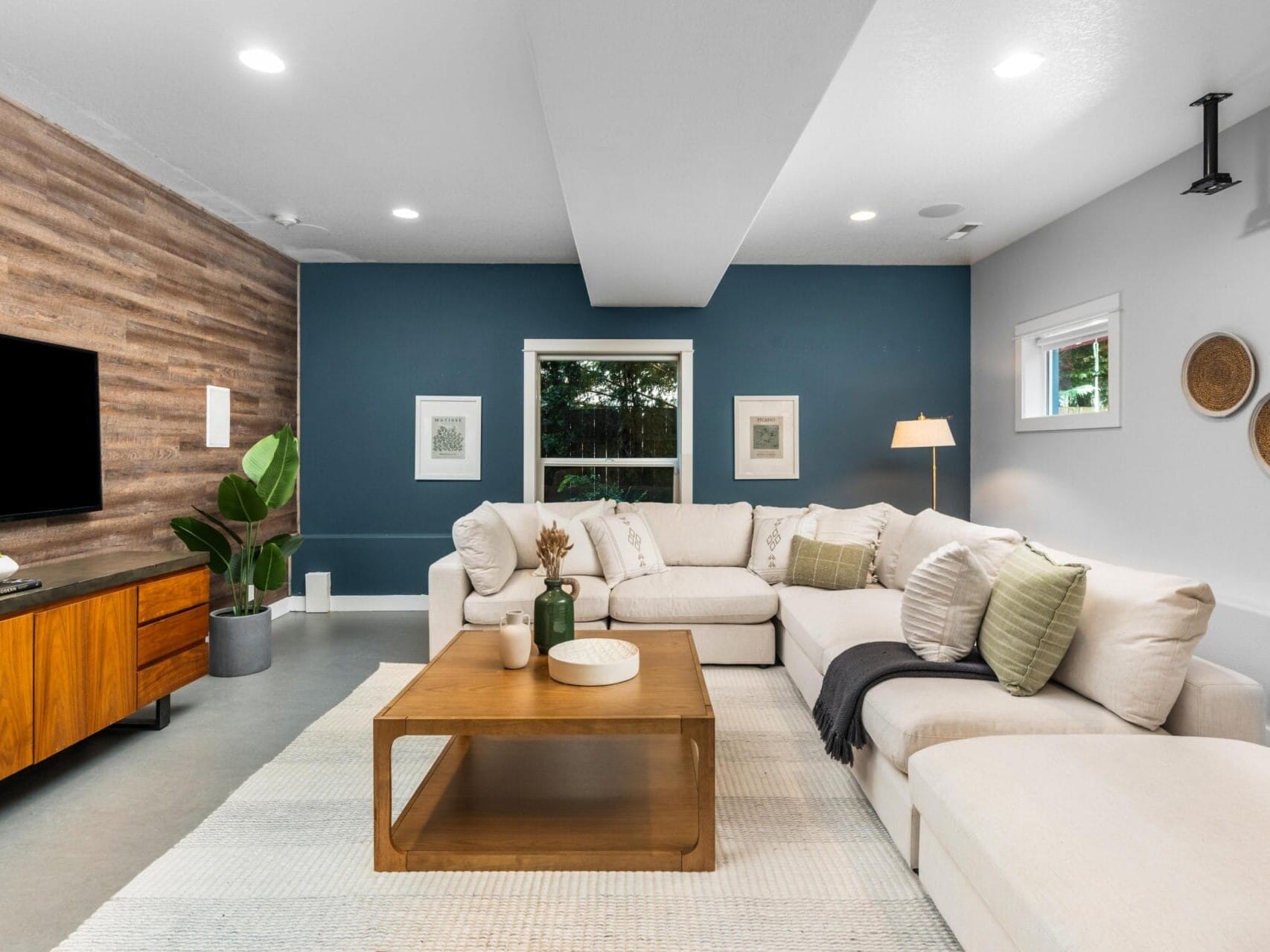 A modern living room with a sectional sofa, wood coffee table, TV on a wooden cabinet, potted plant, and wall art. The walls are blue and white with wood paneling, and a large window lets in natural light.