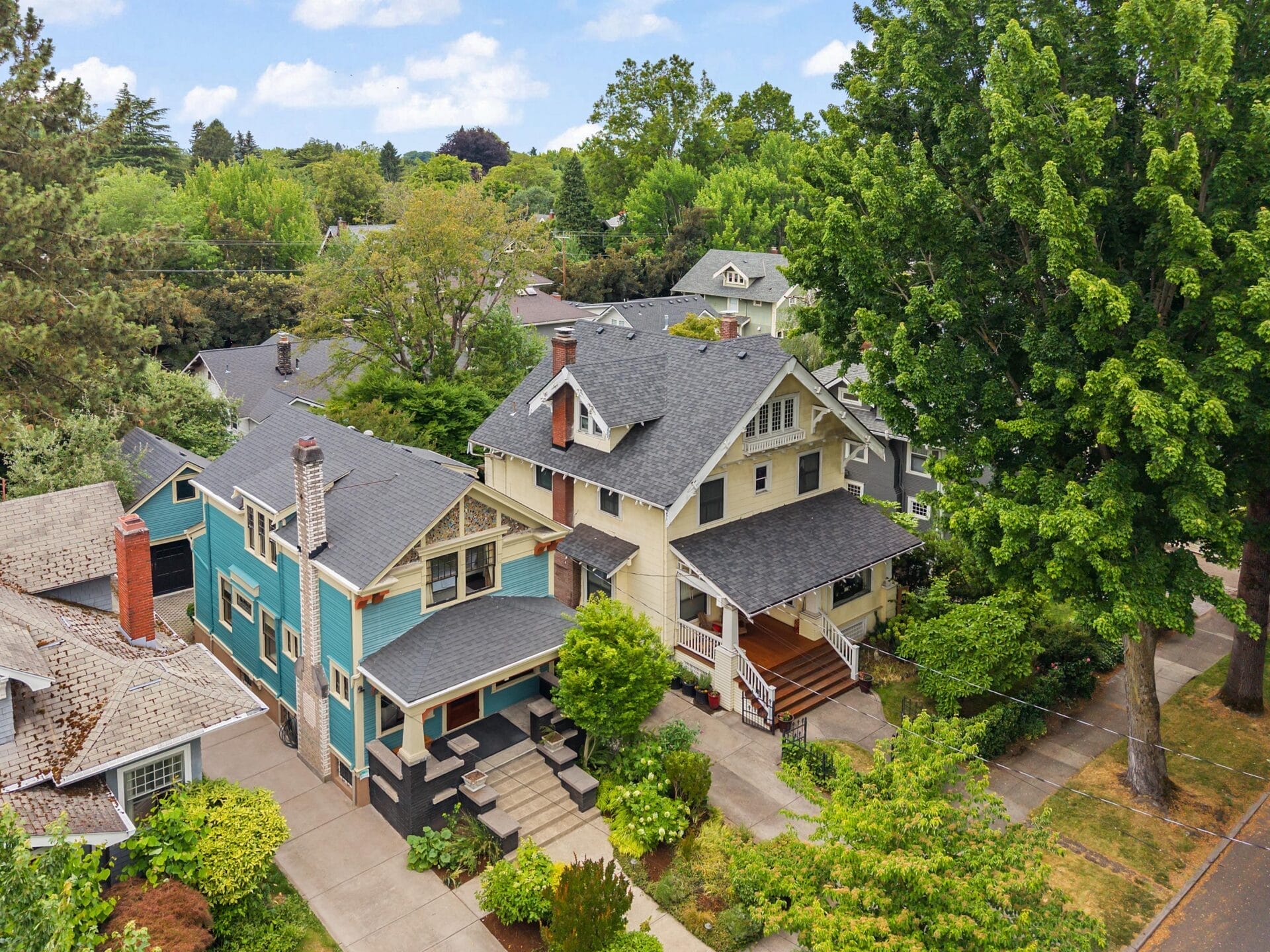 Aerial view of a residential neighborhood with craftsman-style houses, mature trees, and green landscaped yards on a sunny day. Two homes are prominent: one pale yellow with a porch and one blue with a brick chimney.