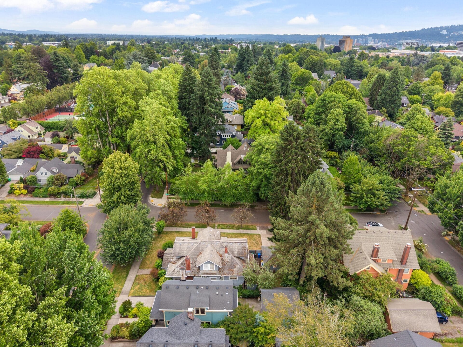 Aerial view of a residential neighborhood with tree-lined streets and houses surrounded by lush green foliage, set against a backdrop of distant city buildings and hills under a partly cloudy sky.