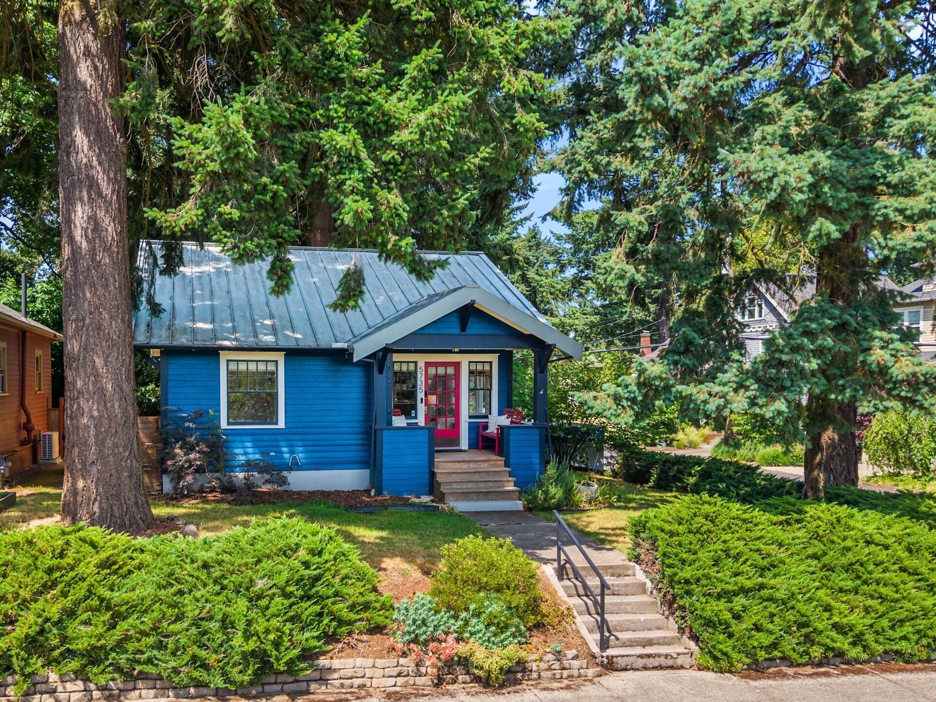 A small blue house with a metal roof and bright red double front doors sits among tall evergreen trees and green shrubs, with a walkway and steps leading up to the porch.