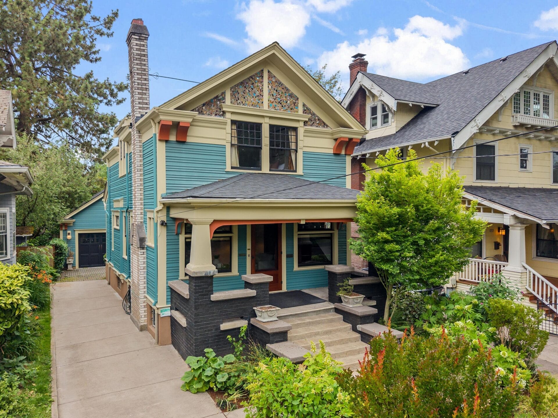 A colorful two-story house with blue siding, beige trim, and stained glass window. It has a front porch, steps, and lush green plants in the yard, with a driveway to the left and neighboring homes visible.
