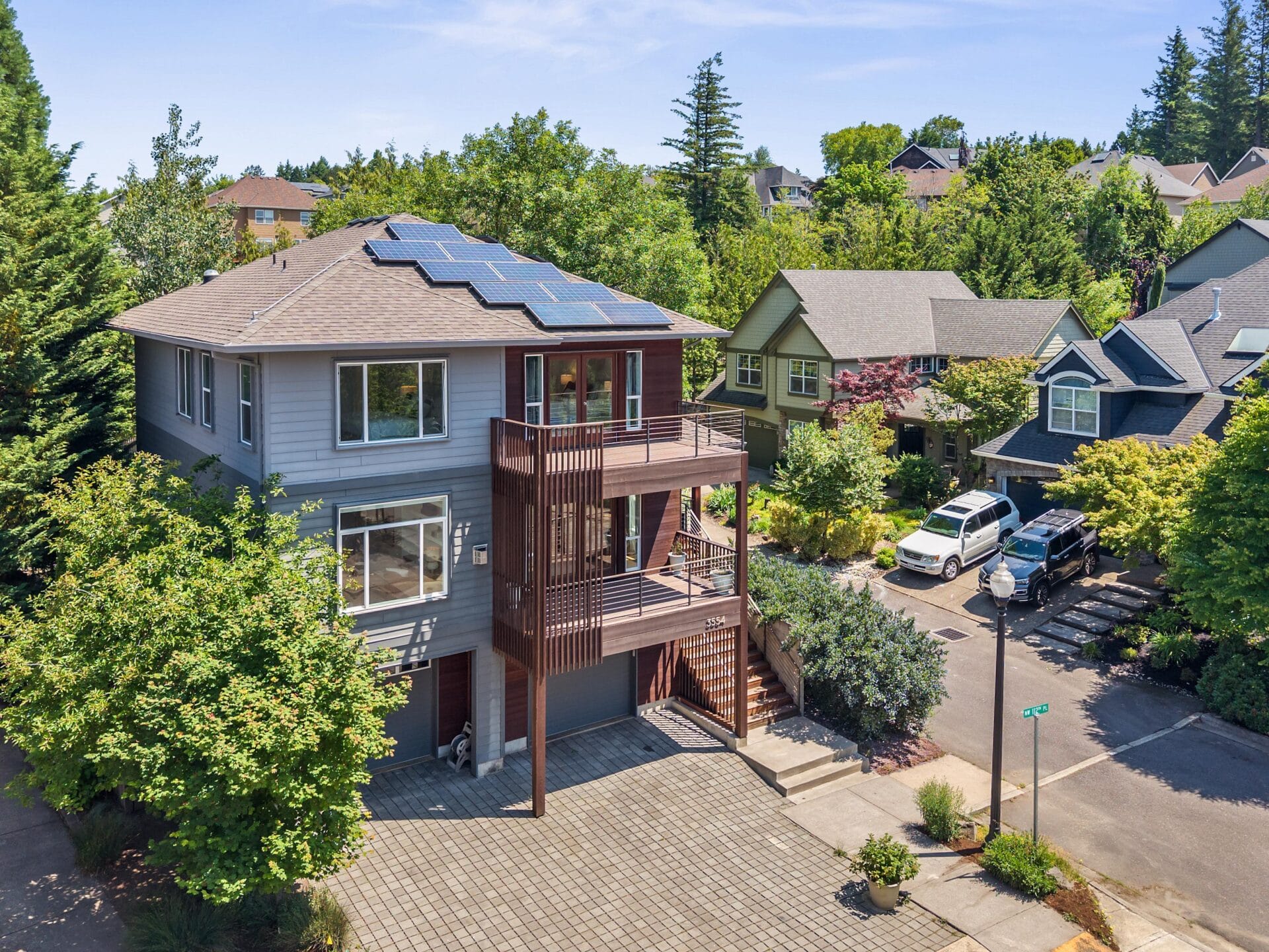 A modern two-story house with solar panels on the roof, large windows, and a wooden balcony, surrounded by trees and neighboring homes on a sunny day. Several cars are parked on the quiet residential street.