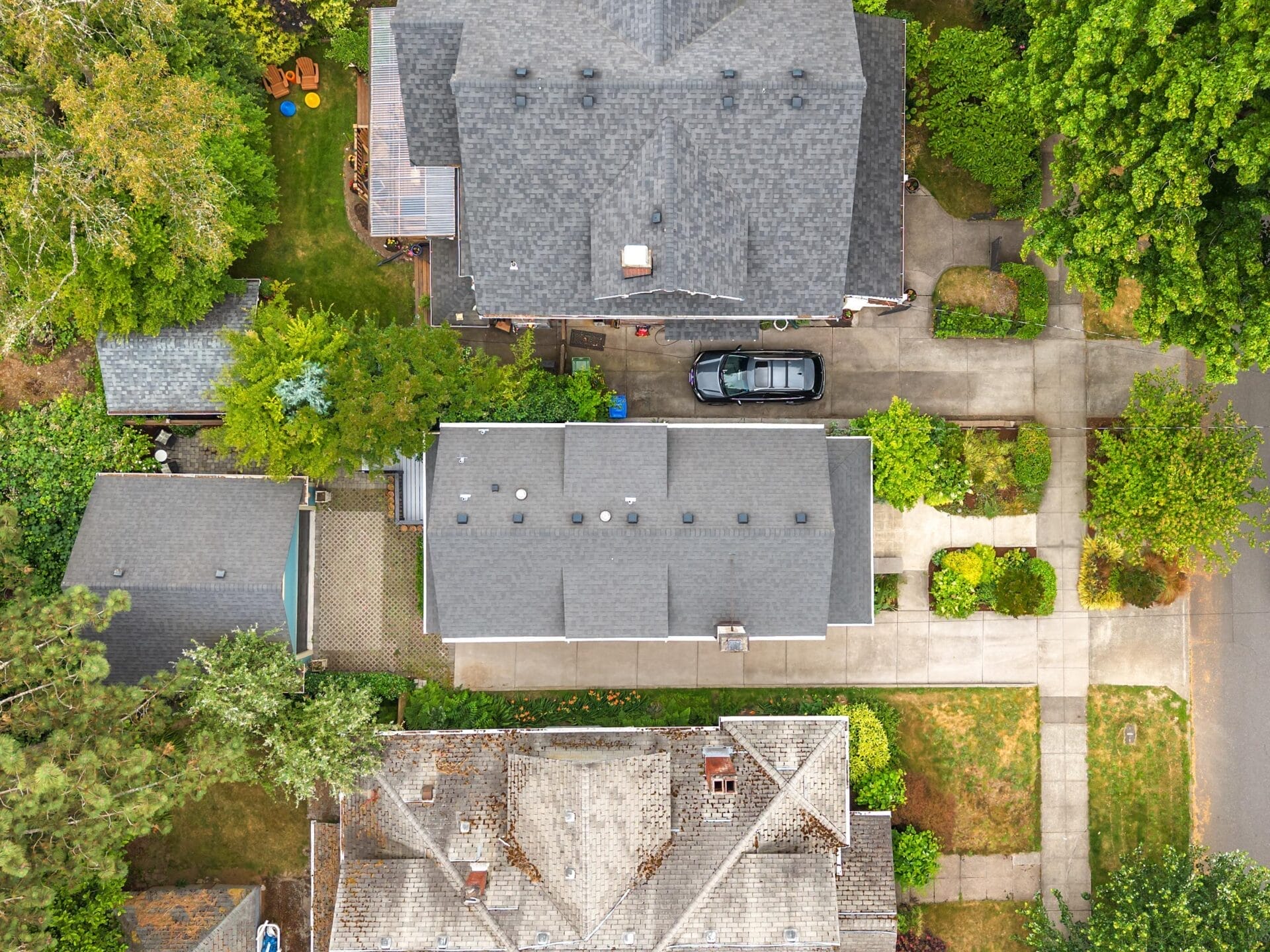 Aerial view of suburban houses with gray rooftops, driveways, a black car parked between two homes, green trees, and neatly landscaped gardens.