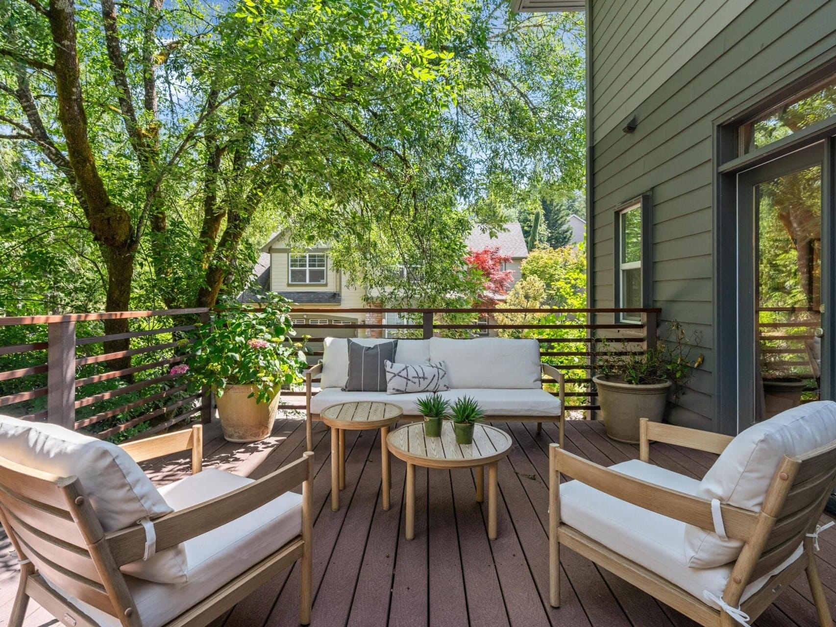 A cozy outdoor patio with cushioned chairs and a sofa around small round tables, set on a wooden deck surrounded by green trees and potted plants, next to a modern house with large windows.