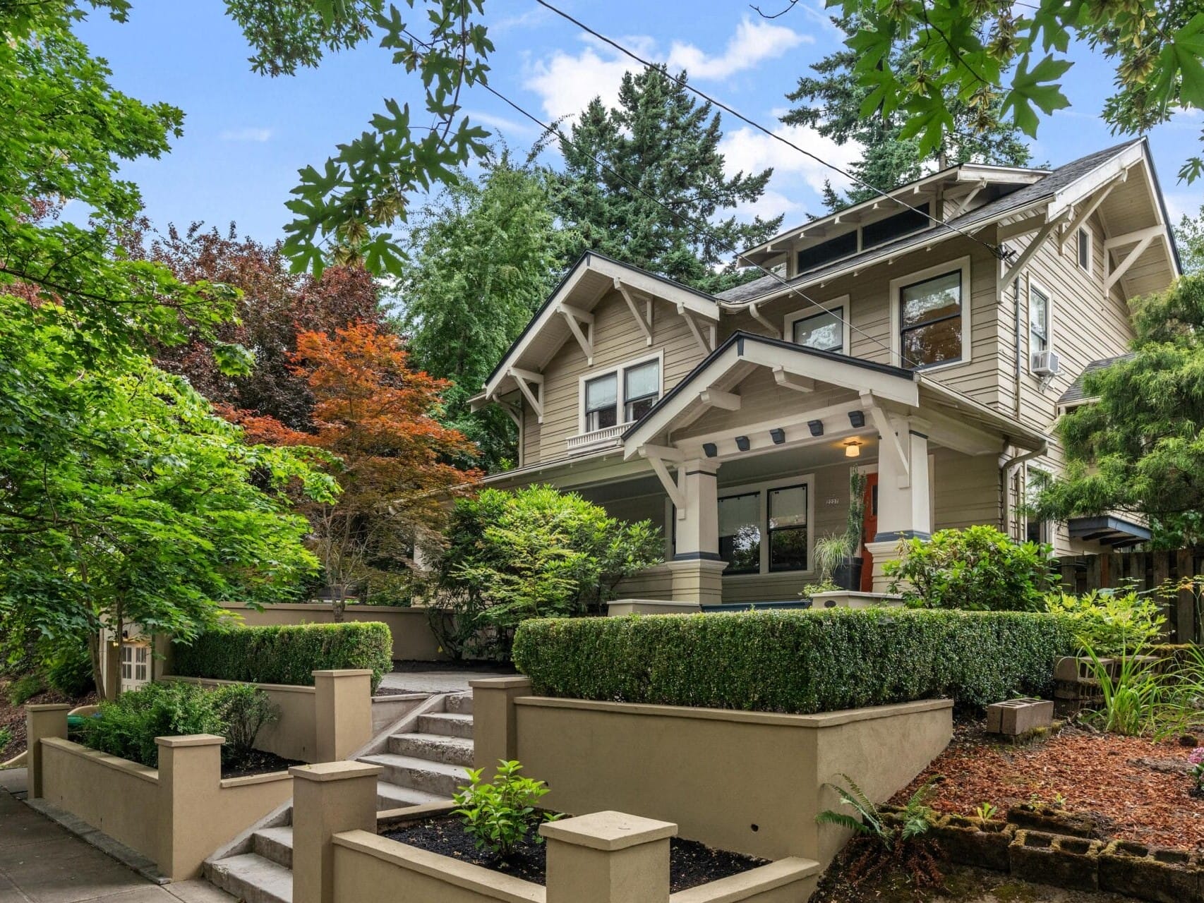A two-story Craftsman-style house with a wide front porch, surrounded by lush green trees and landscaped bushes, with steps leading up to the entrance and a manicured front yard.