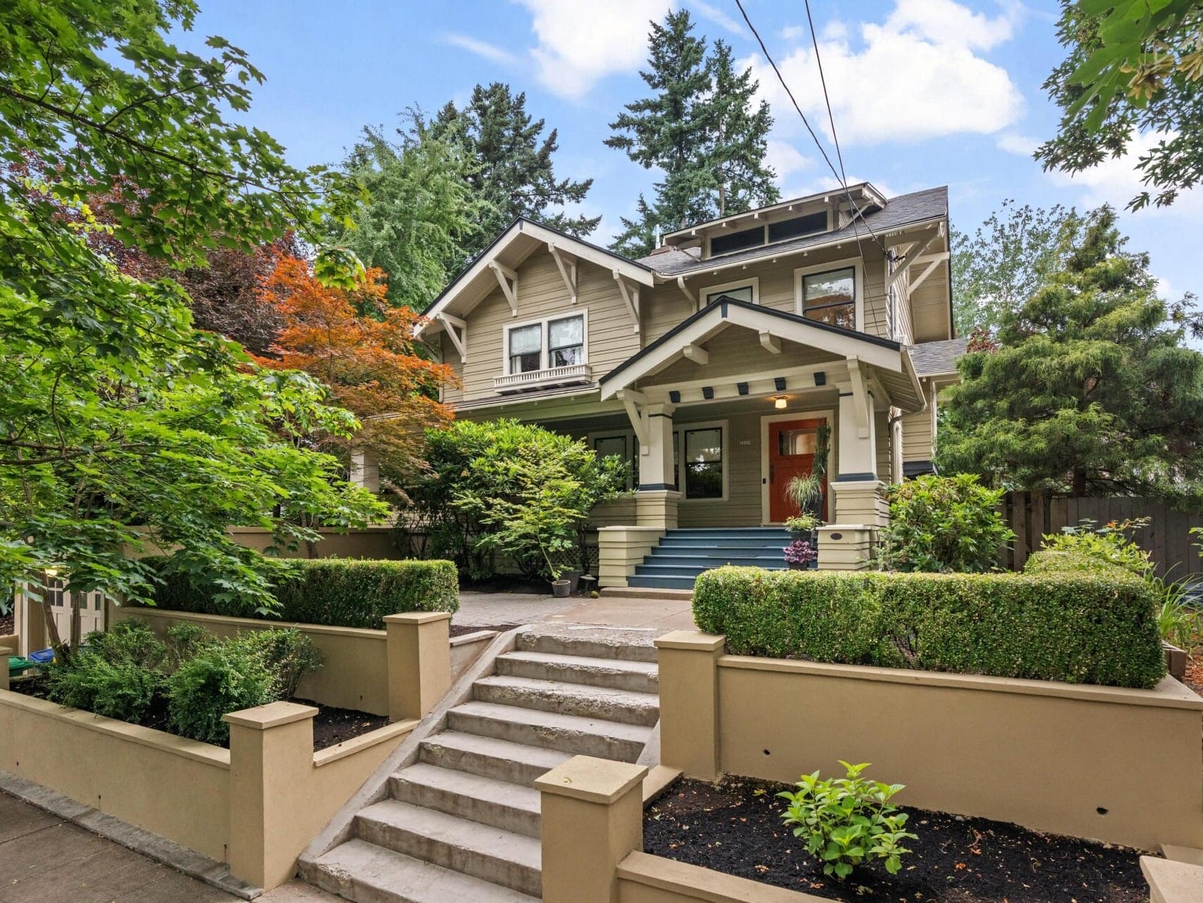 A two-story Craftsman-style house with a covered front porch, beige exterior, and blue steps, surrounded by lush green trees and neatly trimmed hedges. Concrete steps lead up to the entrance.