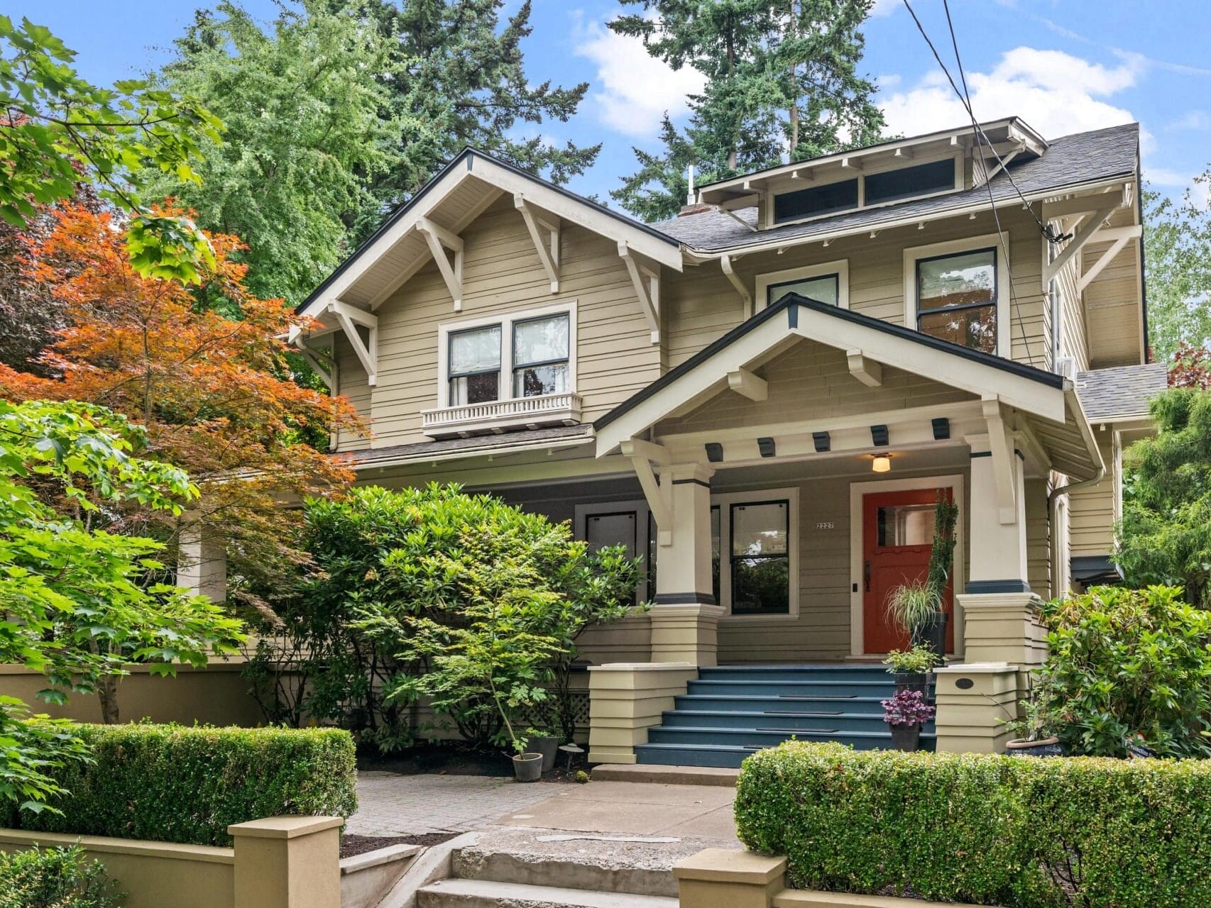 A two-story Craftsman-style house with beige siding, a covered front porch, wide steps, and surrounded by lush green trees and shrubs. The yard features manicured bushes and a concrete walkway.