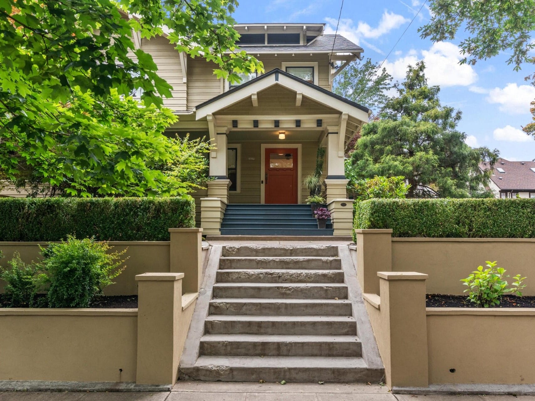 A beige two-story house with a red front door, covered porch, and wide steps leading up through a landscaped front yard with bushes, trees, and a low retaining wall.