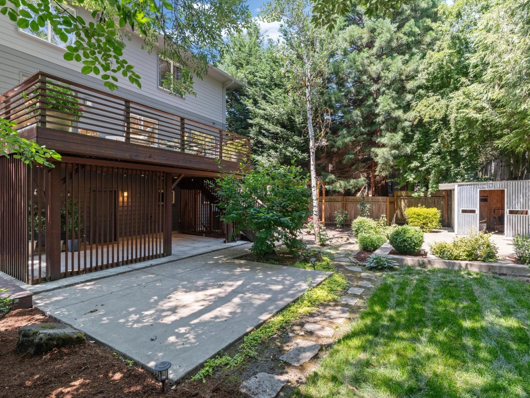 A backyard with a raised wooden deck, sheltered patio below, green grass, shrubs, stone path, tall trees, and a small shed or chicken coop near a wooden fence.