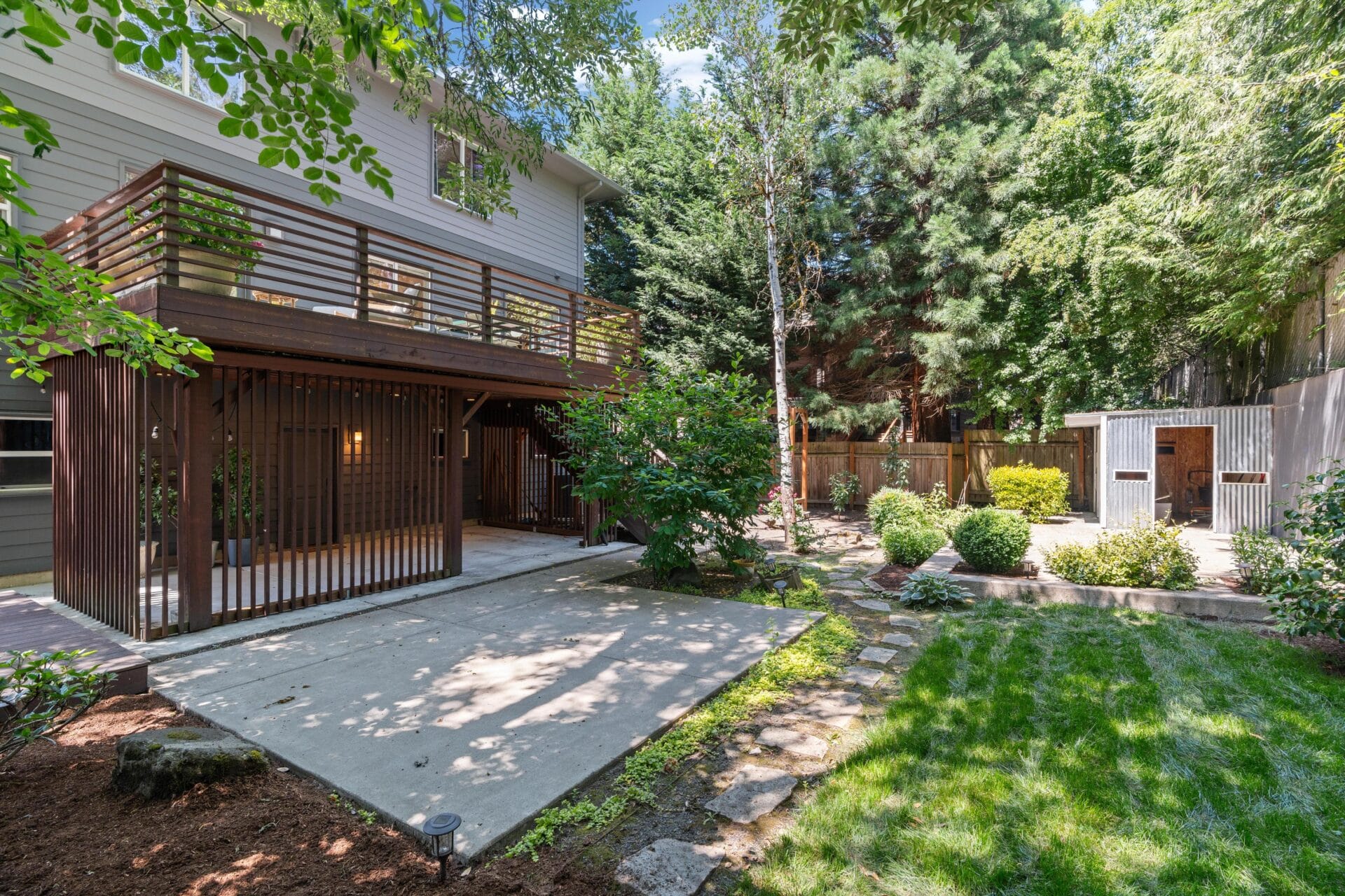 A backyard with a raised wooden deck, sheltered patio below, green grass, shrubs, stone path, tall trees, and a small shed or chicken coop near a wooden fence.