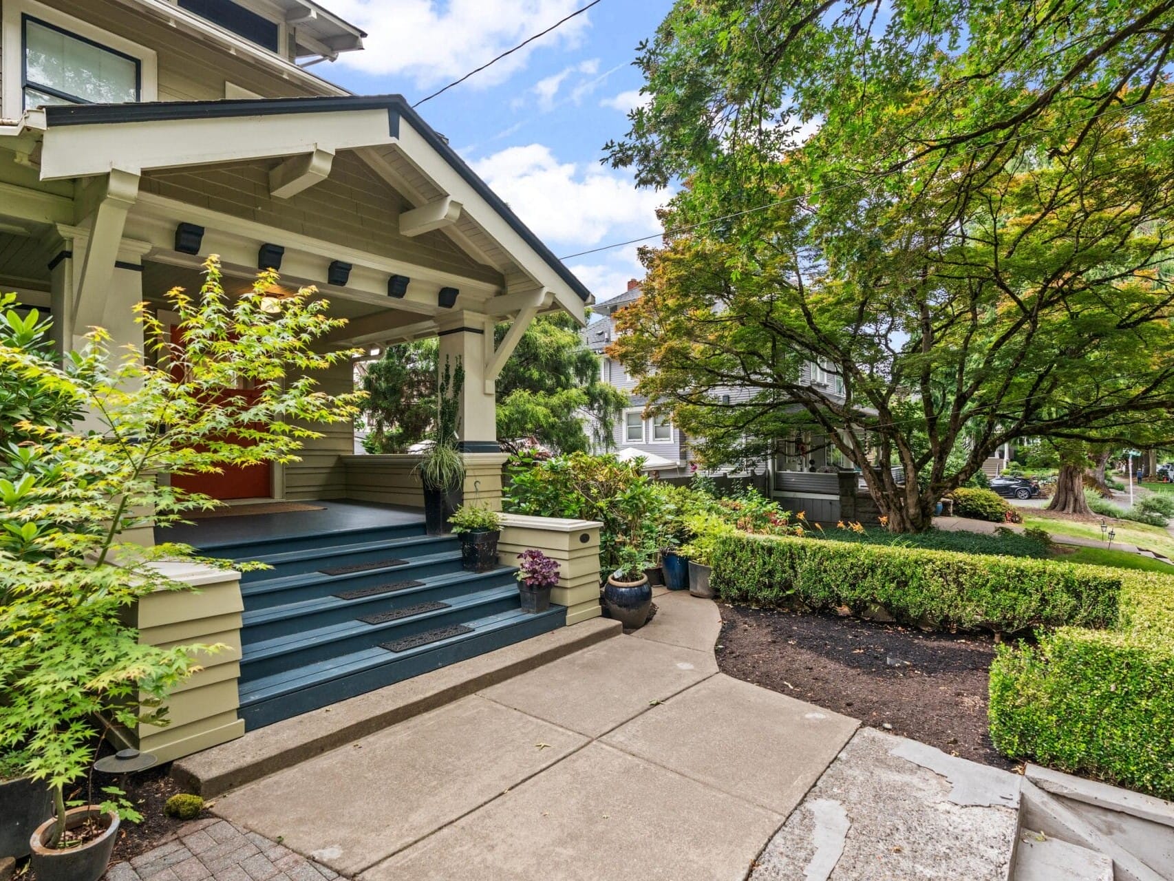 A charming house with a covered front porch, blue steps, potted plants, and lush greenery. A sidewalk and neatly trimmed hedges line the landscaped front yard on a sunny day.