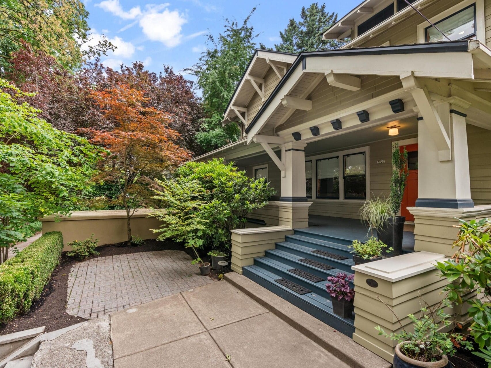 A craftsman-style house with cream siding, blue steps, and an orange front door, surrounded by lush green trees and plants. The entryway features a covered porch and decorative columns.