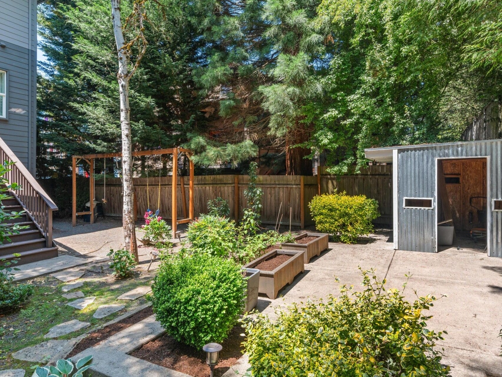 A backyard garden featuring raised planter boxes, lush green shrubs, a modern metal shed, a wooden pergola, a wooden fence, and tall trees providing shade. A staircase leads down from a house on the left.