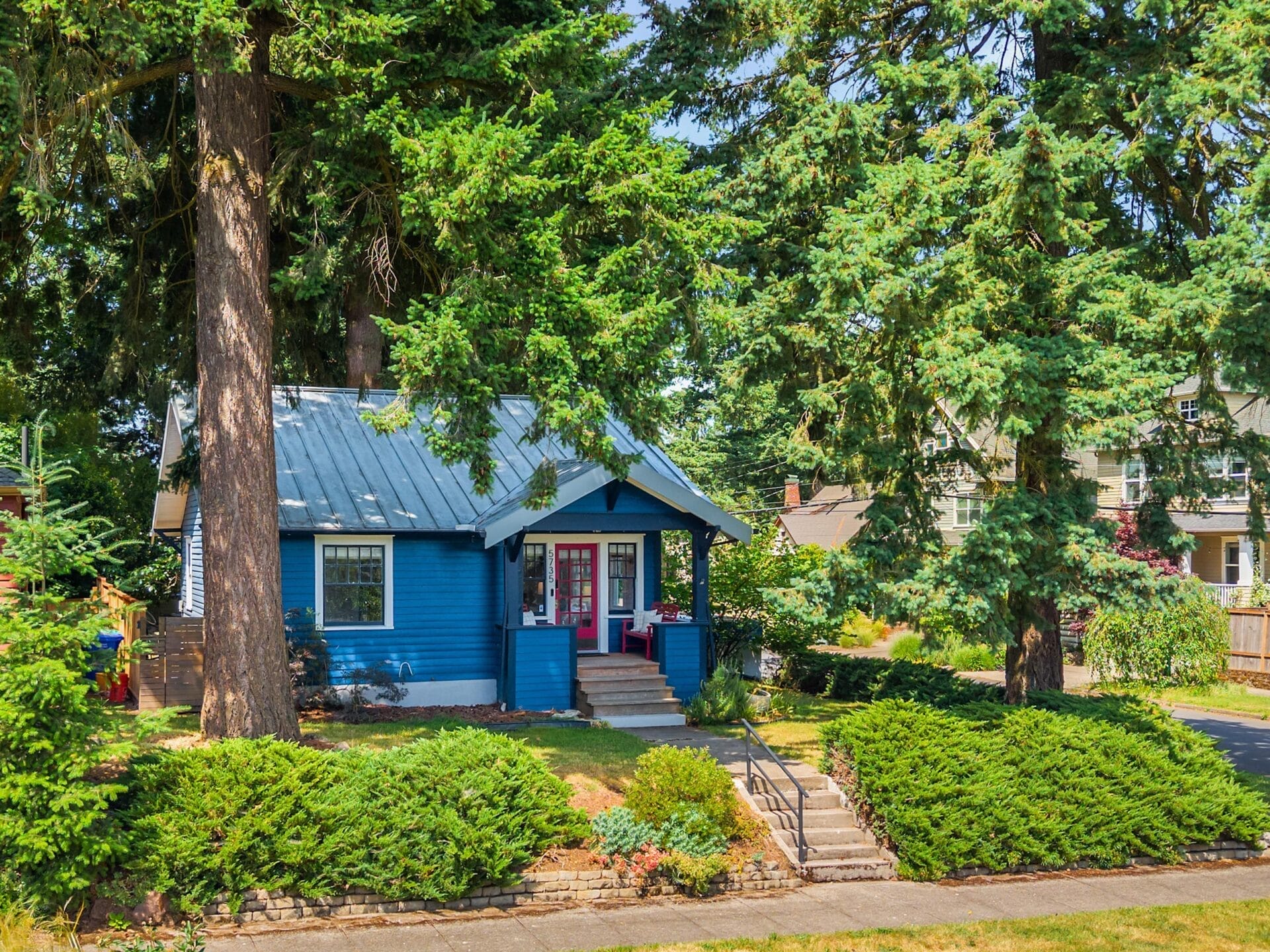 A small blue house with a metal roof and red front door sits among tall evergreen trees and lush green bushes, with a set of steps leading to the porch from the sidewalk.