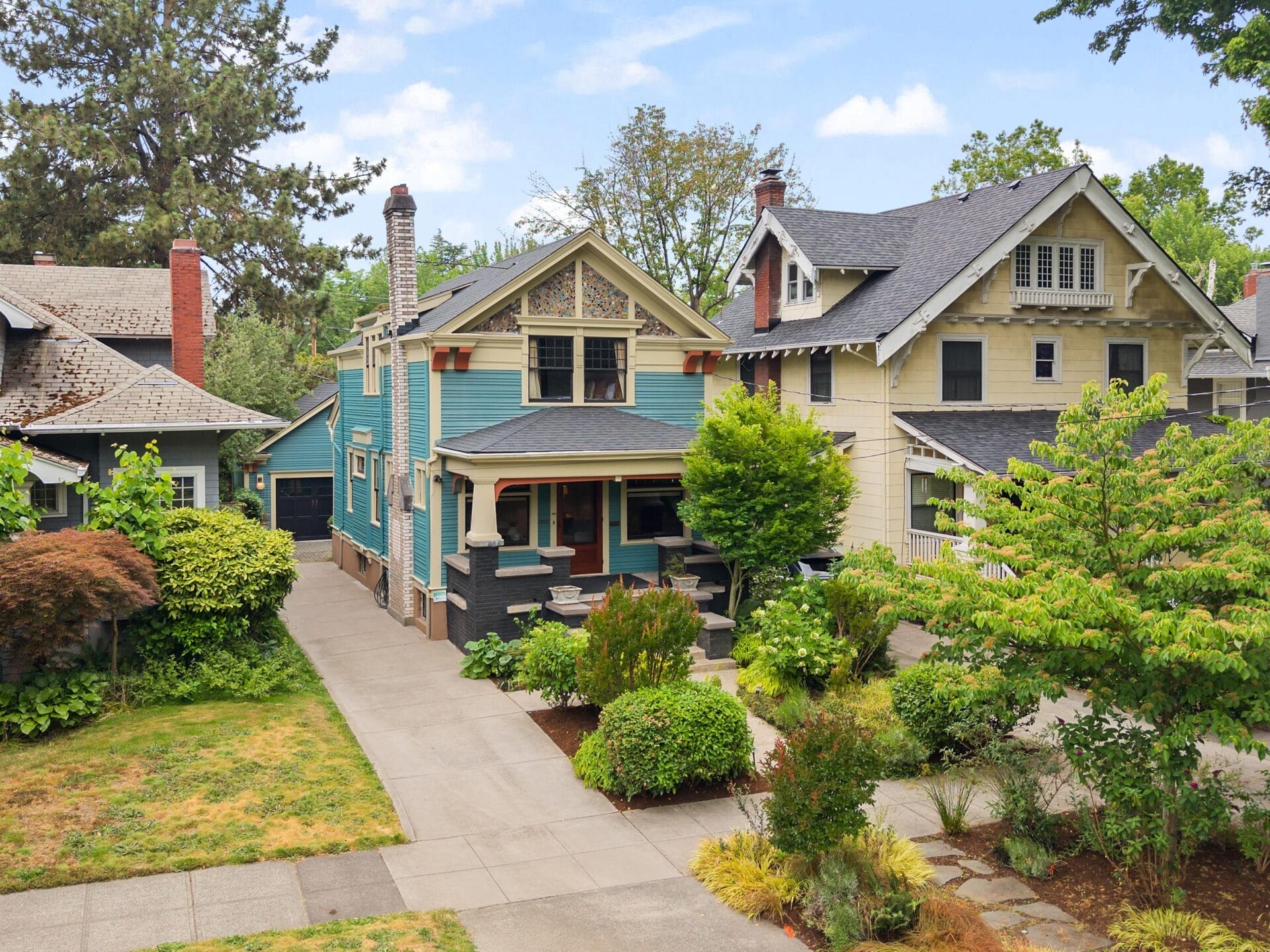 A charming blue and gray two-story house with a covered front porch, surrounded by greenery, sits between similar historic homes on a tree-lined residential street.