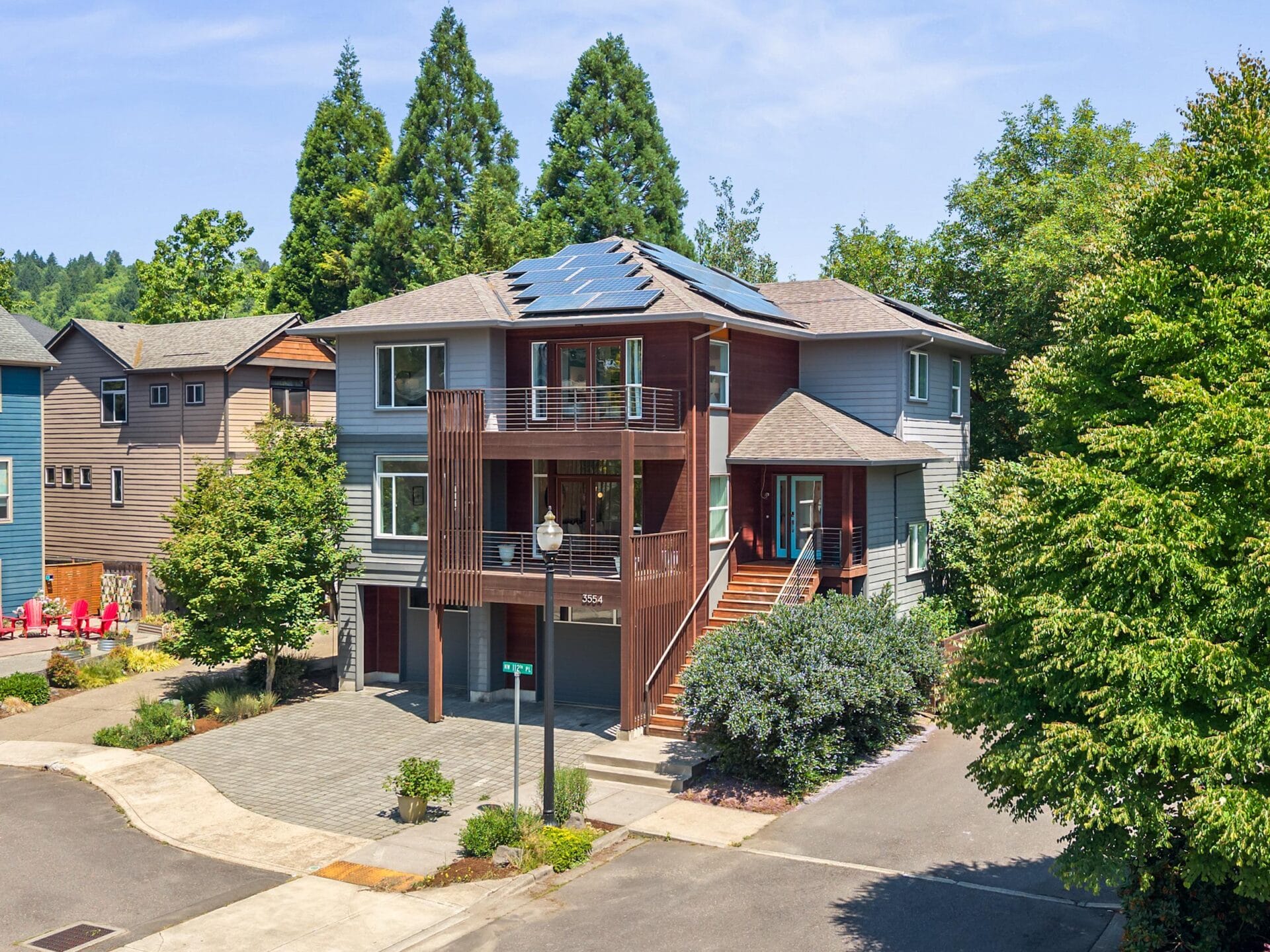 A modern two-story house with blue and wood exterior, a balcony, large windows, rooftop solar panels, and surrounded by trees. The home is located on a corner lot in a suburban neighborhood.