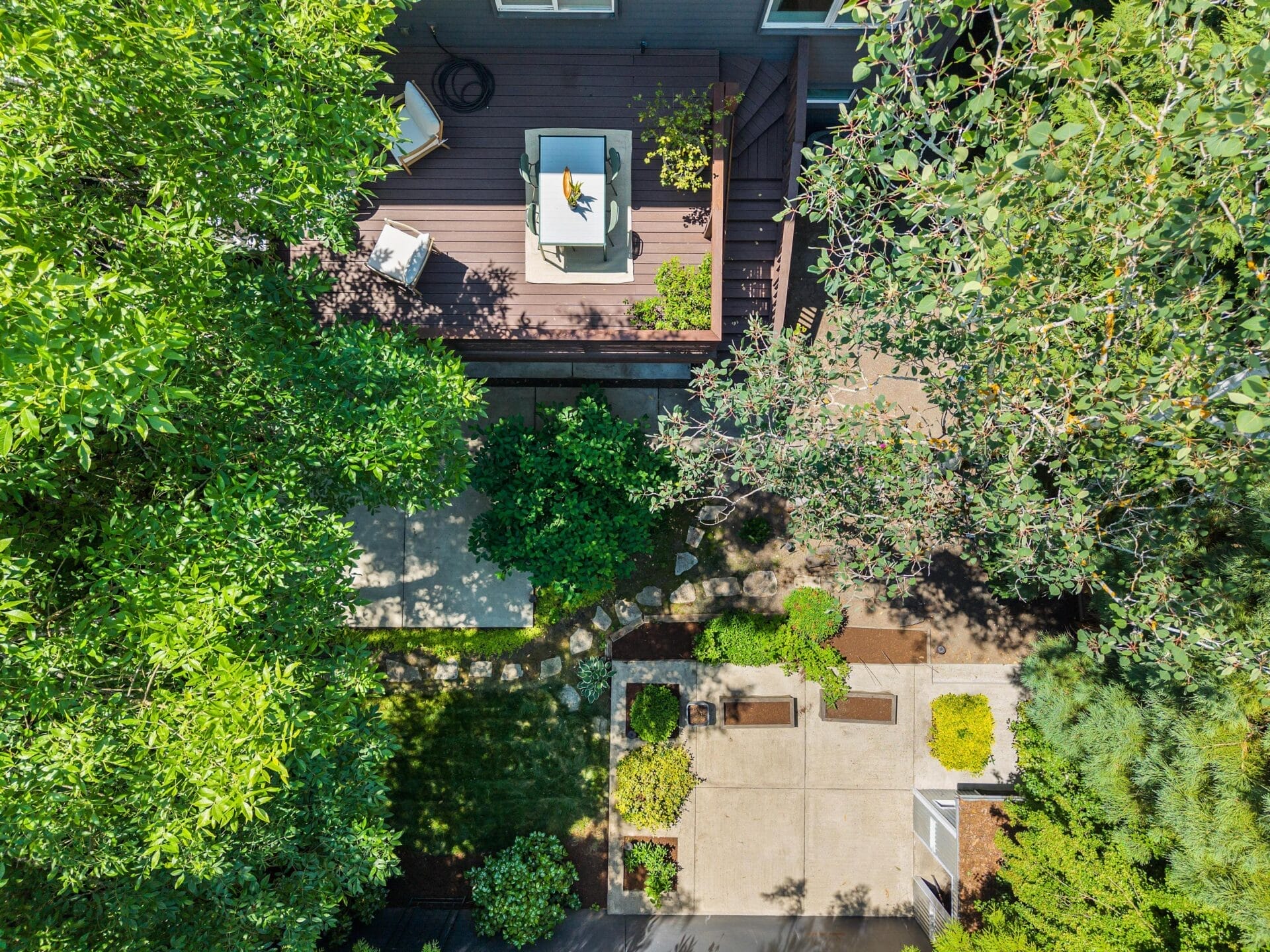 Aerial view of a backyard with a wooden deck, patio furniture, potted plants, a stone pathway, green shrubs, and trees providing shade around the landscaped garden.
