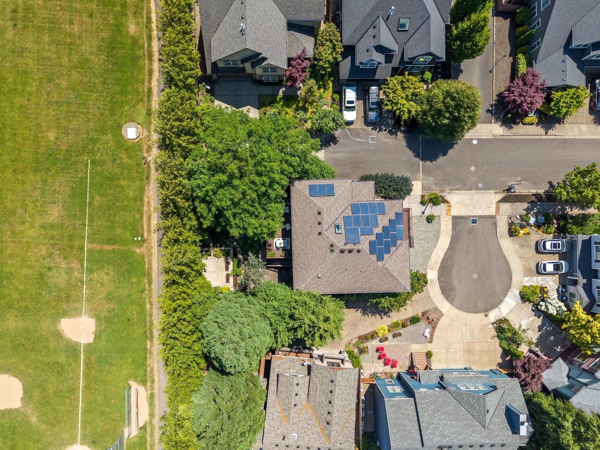 Aerial view of a suburban neighborhood with houses, a cul-de-sac, trees, and a nearby grassy field with baseball diamonds. One house has solar panels on its roof. Cars are parked along the street and driveways.
