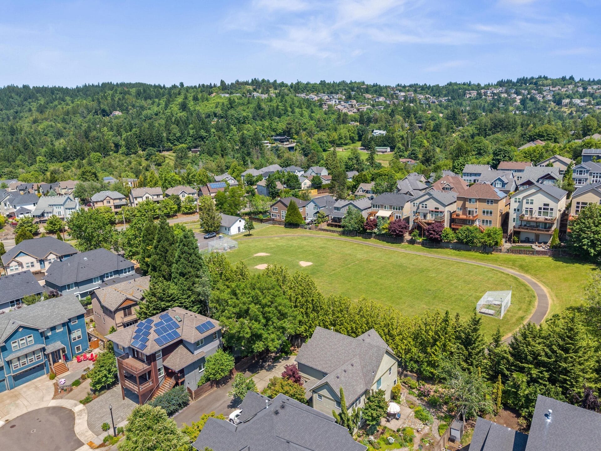Aerial view of a suburban neighborhood with rows of houses, a green baseball field at the center, and tree-covered hills in the background under a partly cloudy sky.